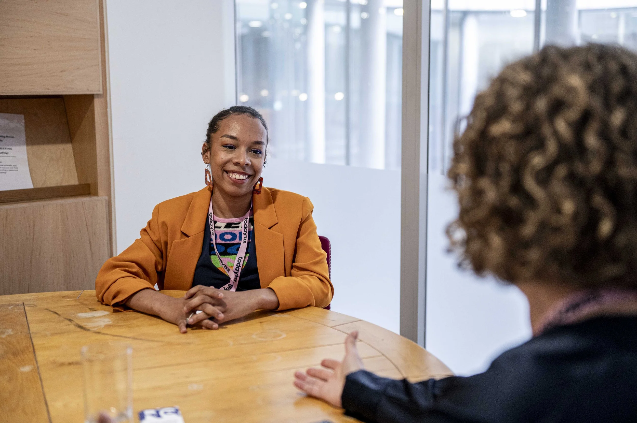 Two women sitting at a wooden table engaged in conversation, one smiling and the other gesturing with her hand, in a modern office setting.