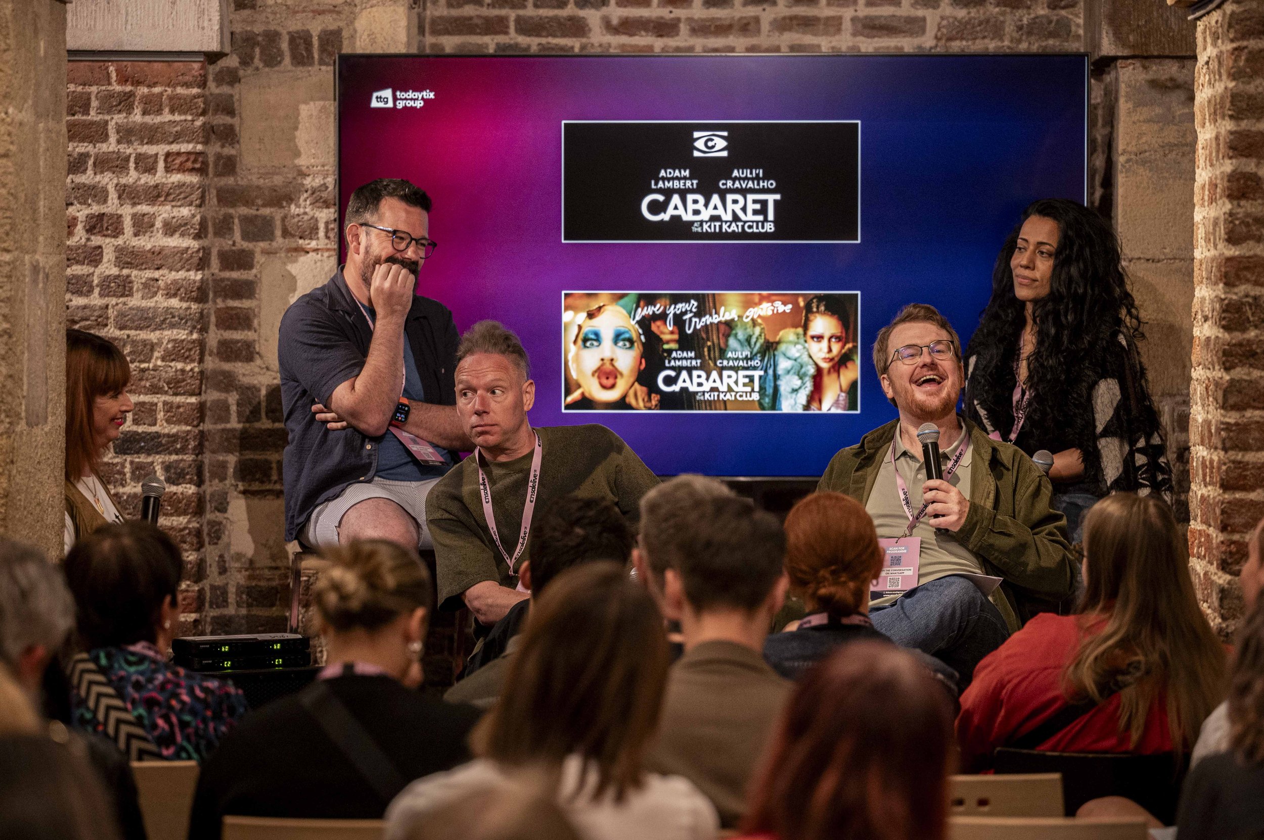 A panel discussion at a conference with four people on stage and a large screen behind them showing promotional material for a cabaret show. Audience members seated and listening.