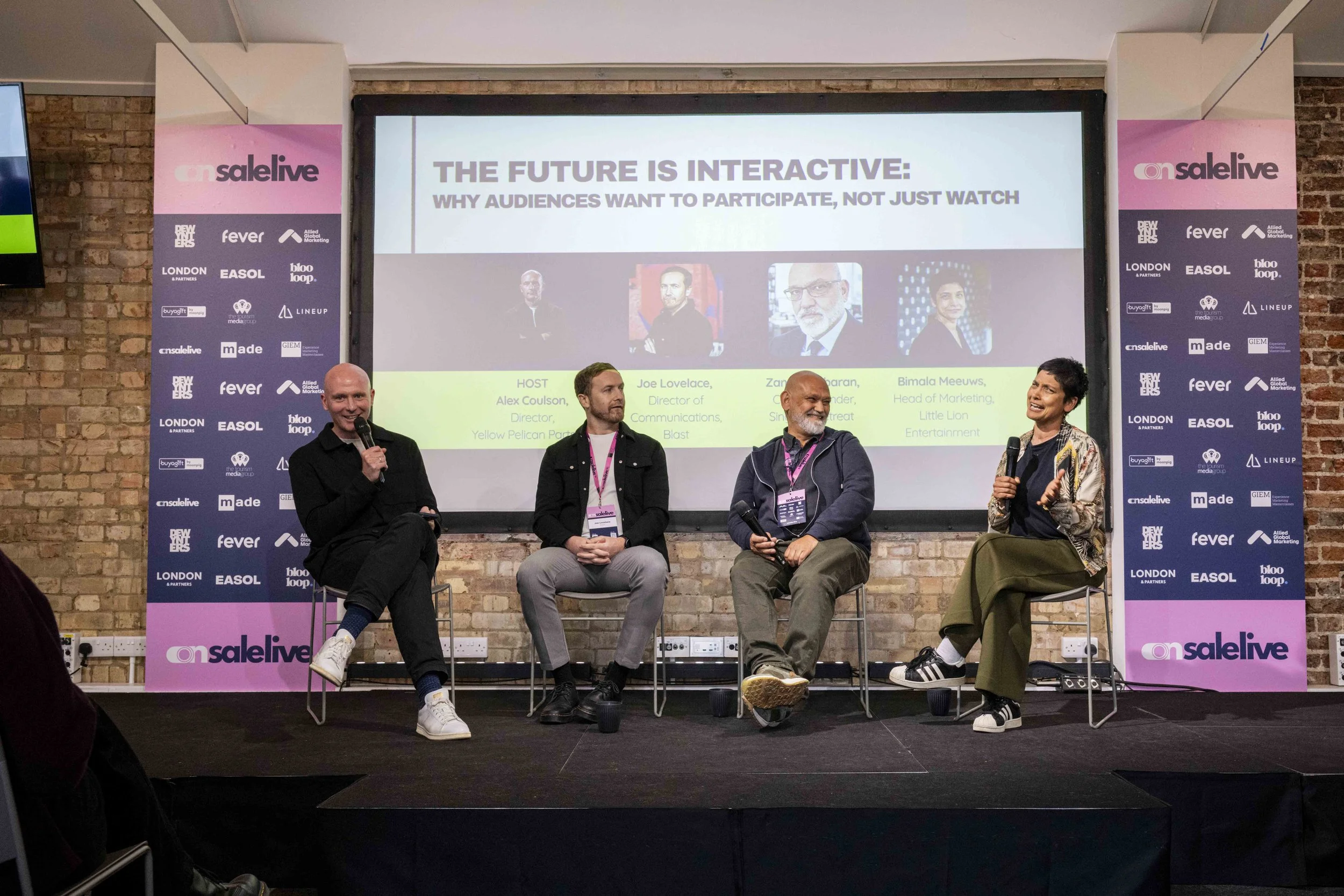 Four people seated on a stage in front of a large screen displaying a presentation titled 'The Future is Interactive: Why Audiences Want to Participate, Not Just Watch'. The stage has pink banners with logos and text, and the background features an e