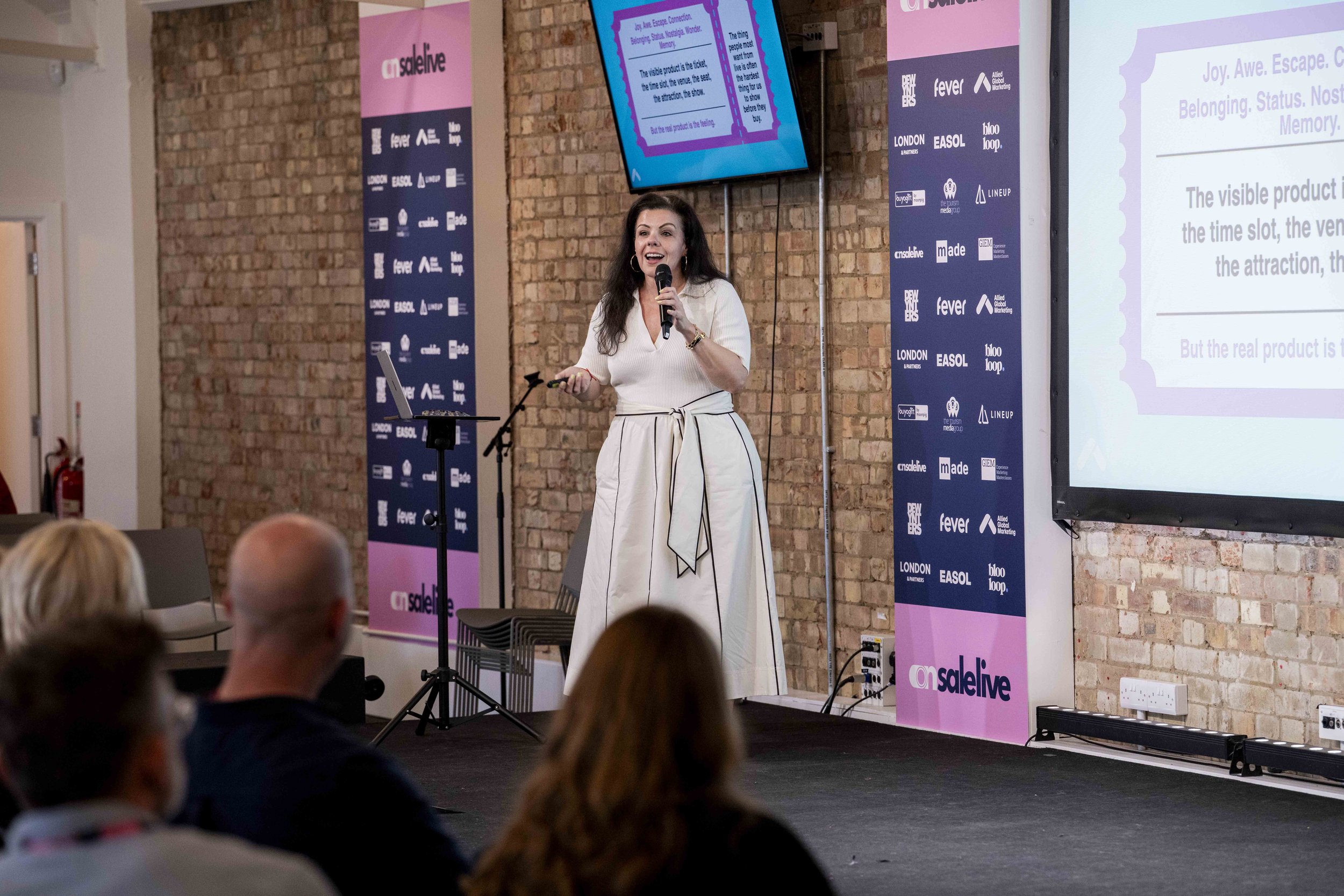 A woman presenter standing on a stage with a microphone in hand, speaking to an audience. She is wearing a white dress with black accents. Behind her are banners with logos and text, and a large screen displaying a presentation slide. The backdrop is a brick wall, and the audience is seated, watching the presentation.