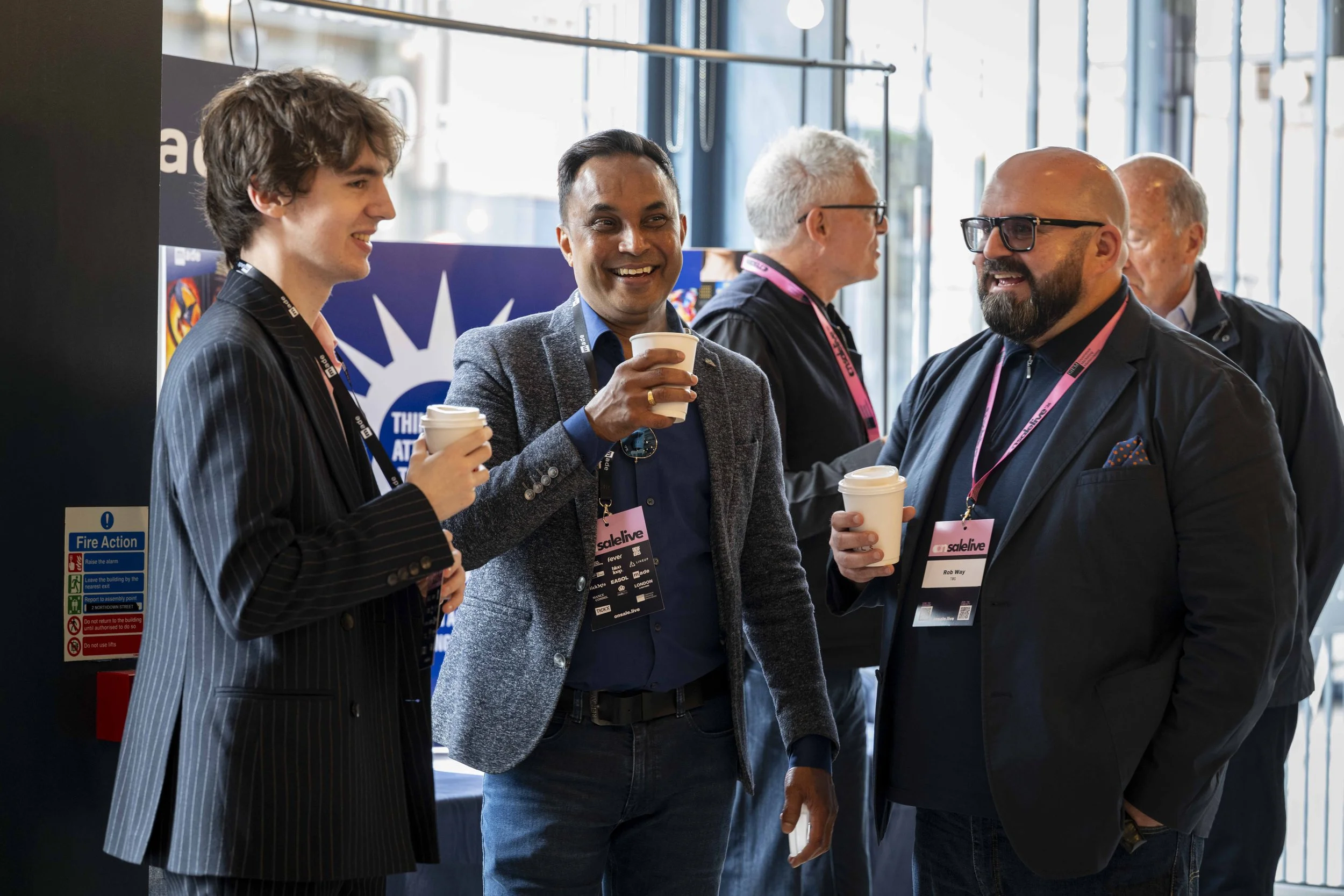 Group of five men standing together at a conference, holding coffee cups and engaging in conversation, with exhibition booths and natural light in the background.