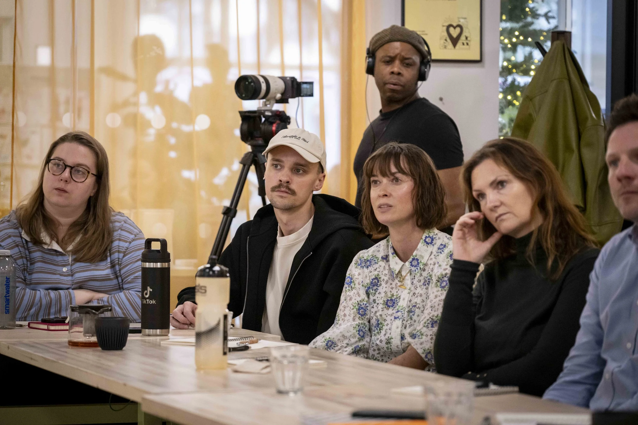 A group of people sitting at a table during a meeting or discussion, with a cameraman filming the event in the background.