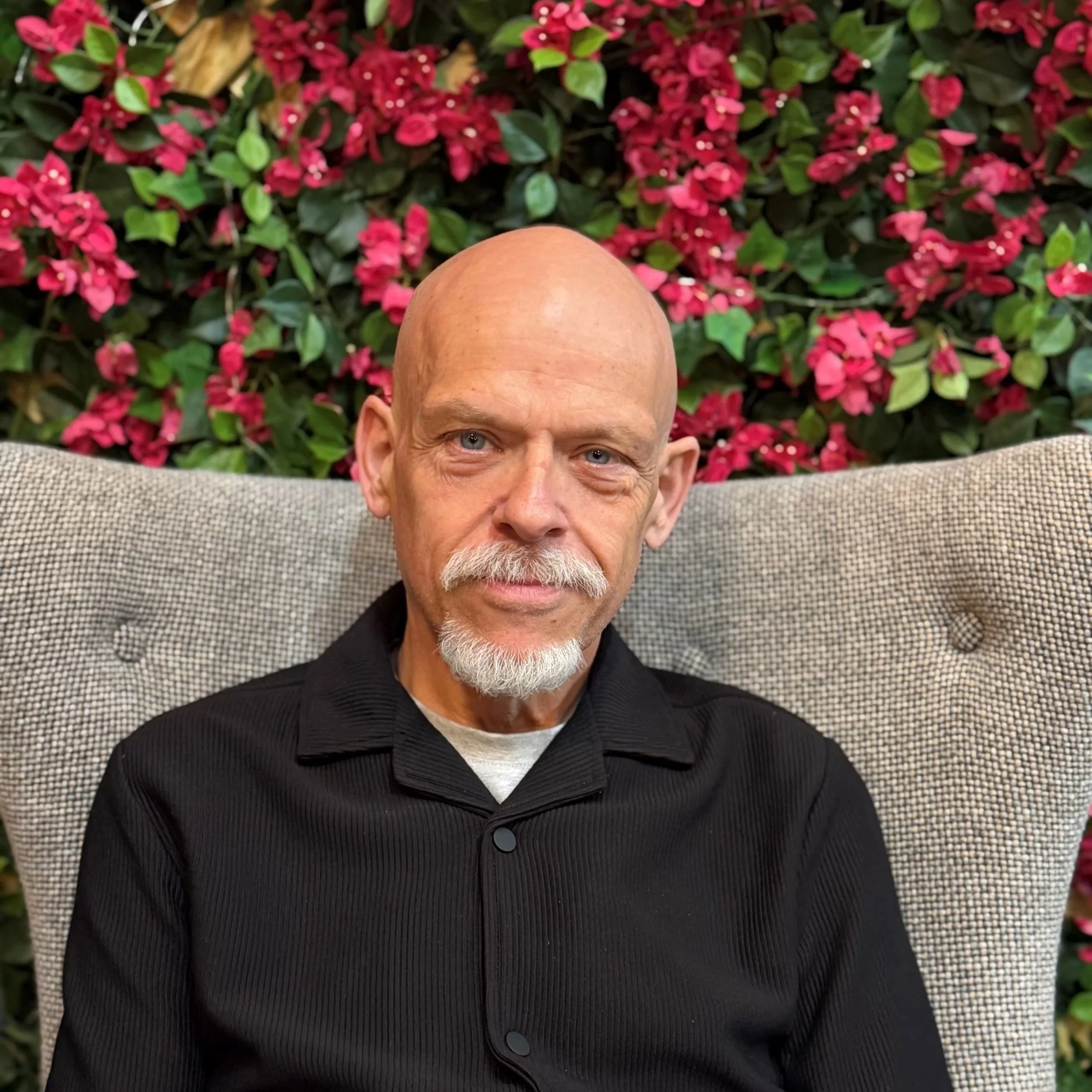 Close-up of a bald man with a white beard, wearing a black shirt, smiling, with pink flowers and green leaves in the background.
