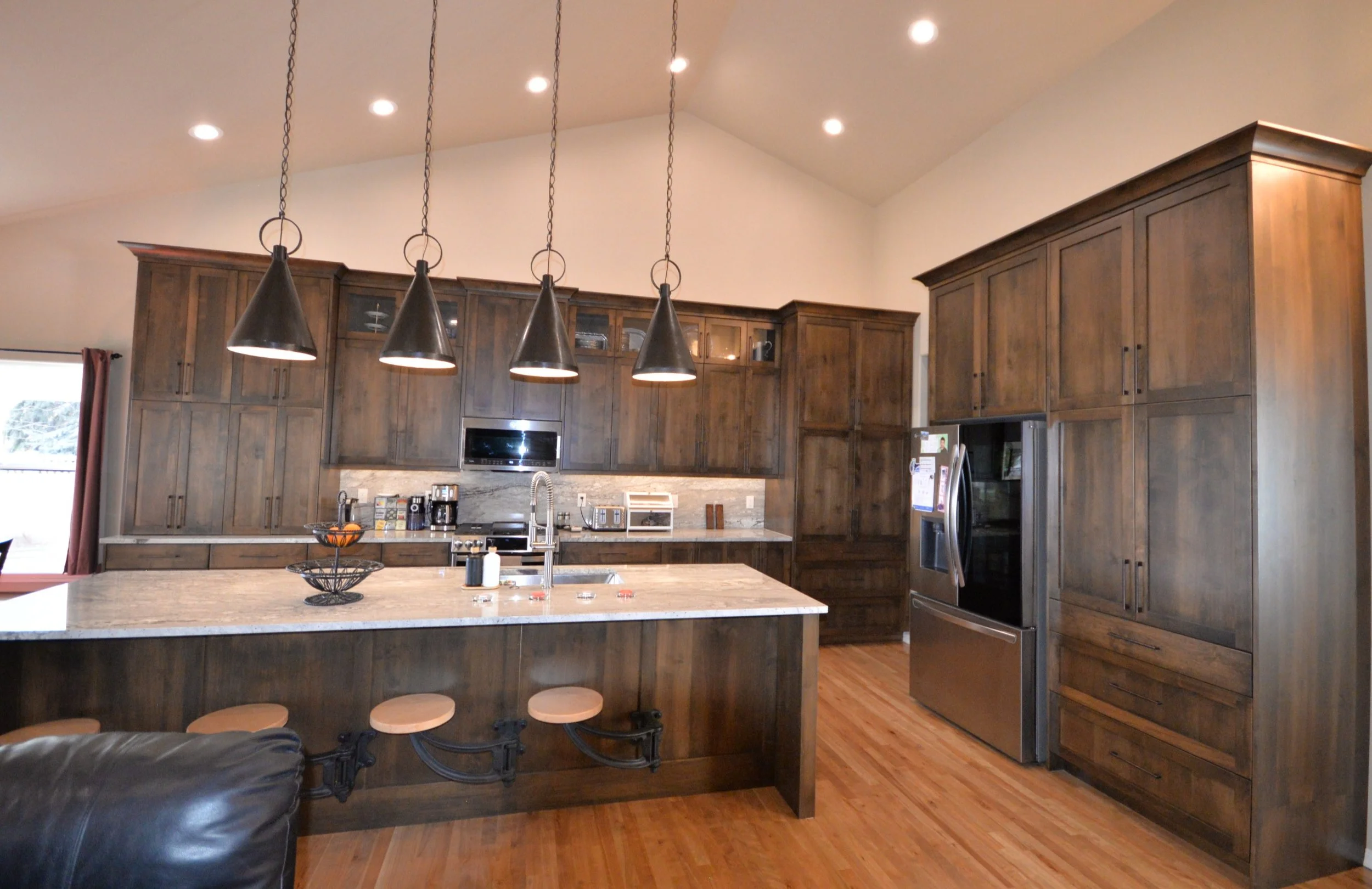 Modern kitchen with stained wood cabinets, quartz countertops, and black pendant lights over a kitchen island with swing away barstools, stainless steel appliances, and hardwood flooring.