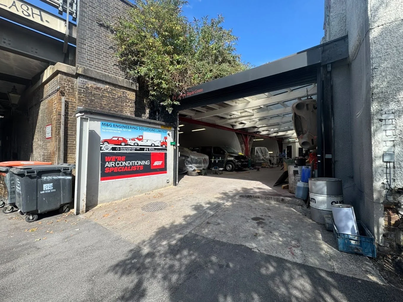An auto repair garage with a partially open garage door, several cars inside, and a sign advertising air conditioning specialists. There are trash bins and some barrels outside.