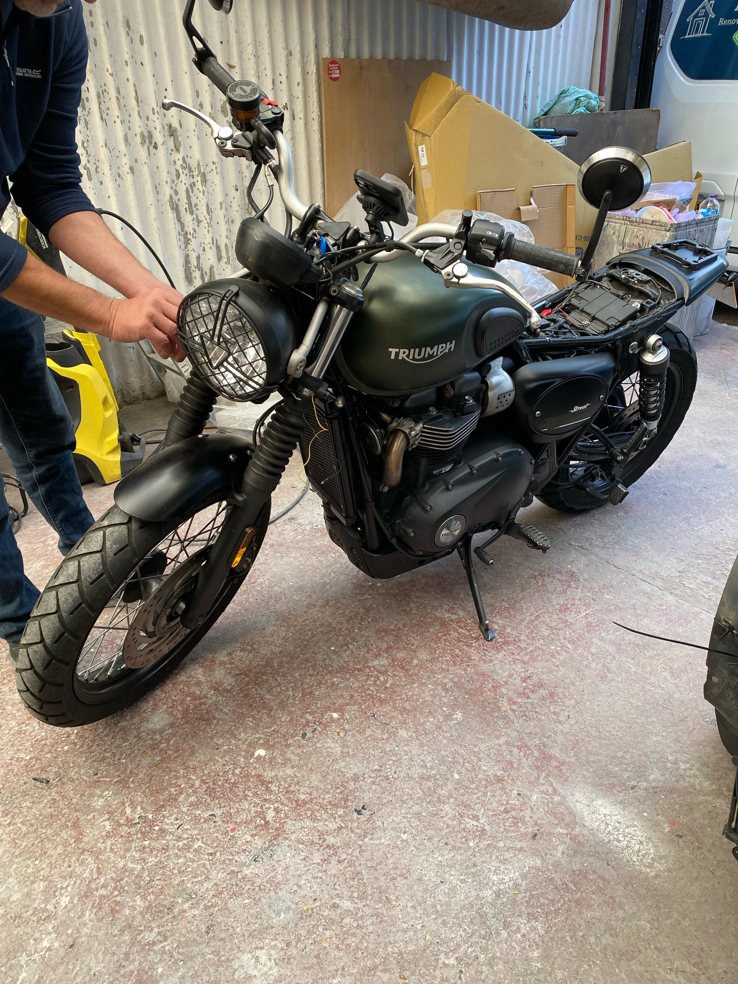 A person working on a partially disassembled Triumph motorcycle in a garage with various boxes and tools in the background.