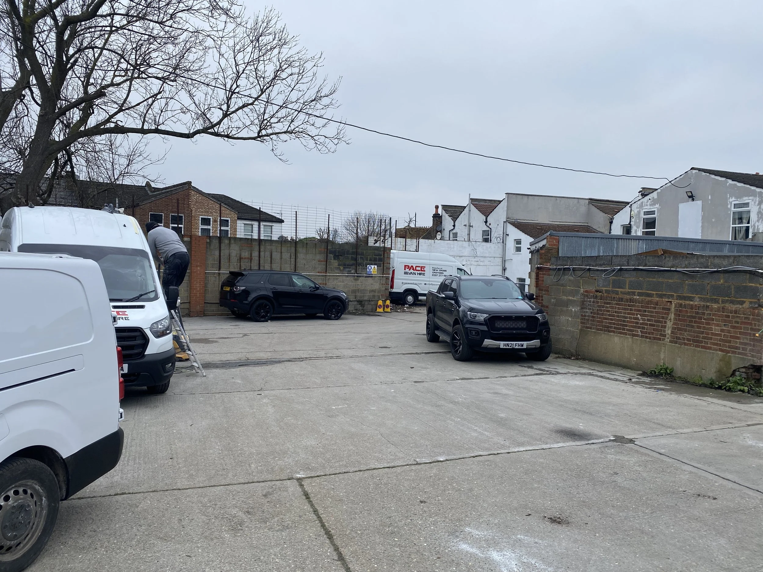 A parking lot with several parked cars, a man working on a vehicle, a white van, a short brick and concrete wall, residential buildings, leafless trees, and a cloudy sky.