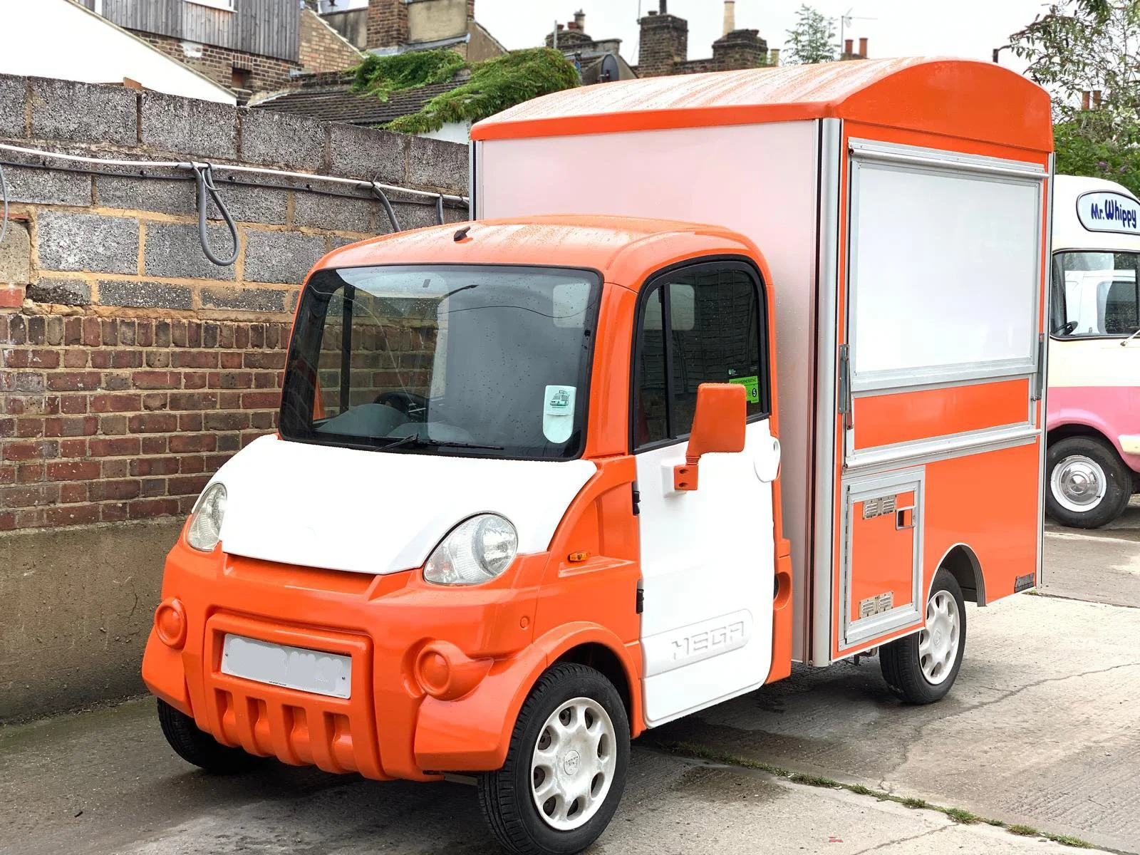 A small orange and white delivery truck parked on a street with a brick wall in the background.