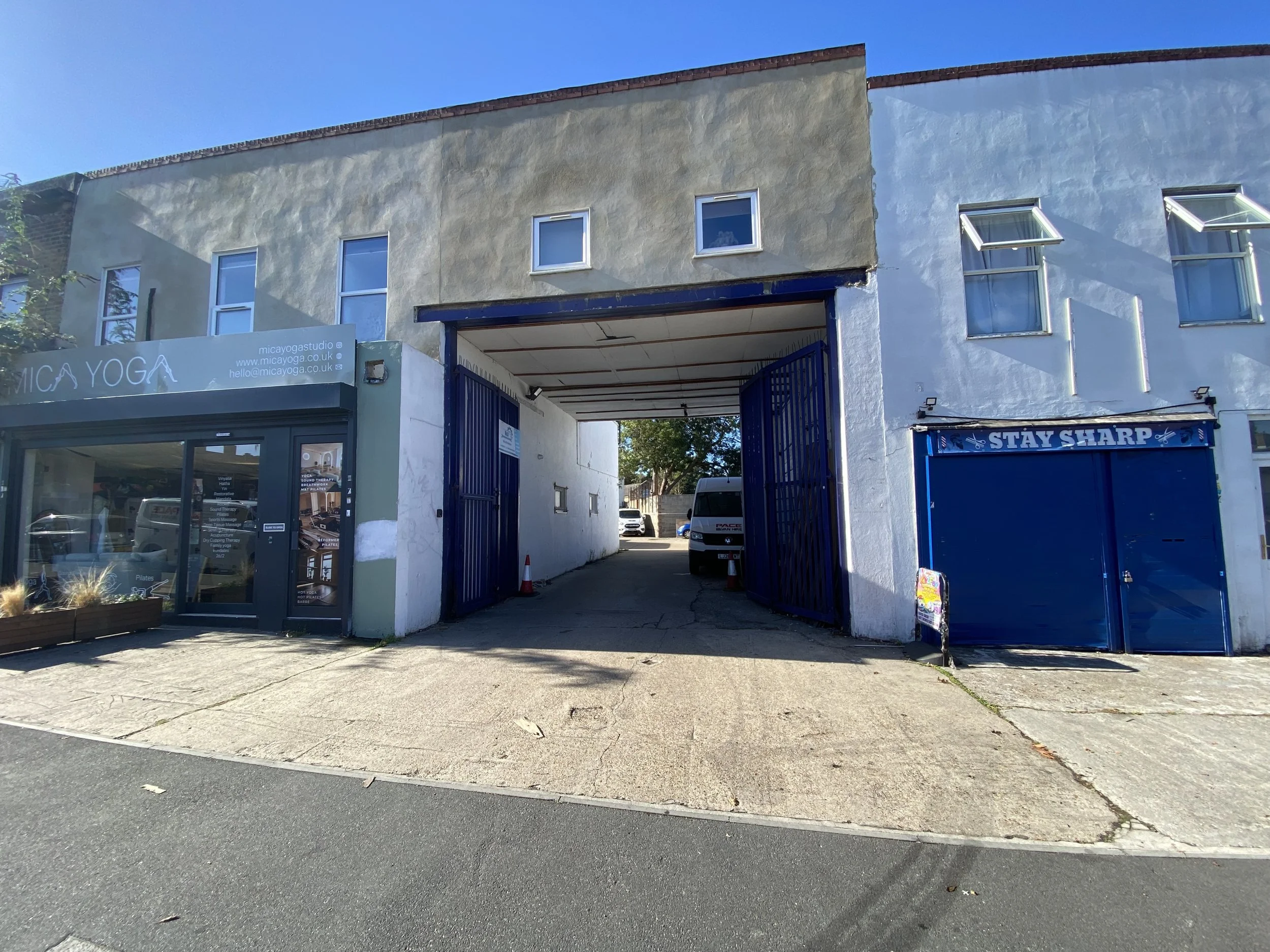 Photo of a building with a yoga studio on the ground floor and an open gate leading into a parking area behind. The building has three windows on the upper floor, with the middle window out of place. There is a blue sign that reads 'STAY SHARP' on the right side of the building.