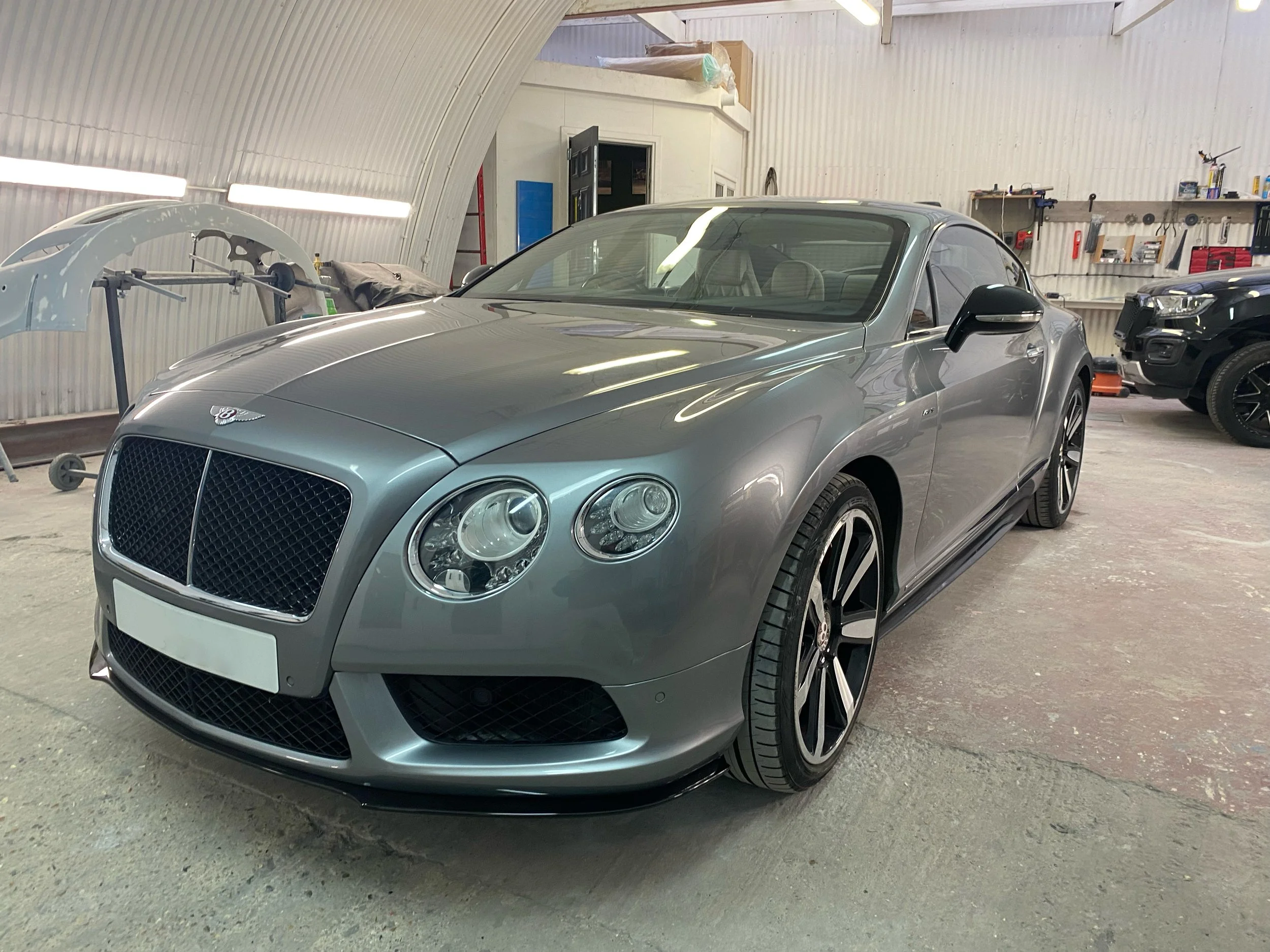 A silver luxury car parked inside a garage with a corrugated metal wall. The garage has tools and equipment on shelves, and another vehicle is partially visible on the right side.