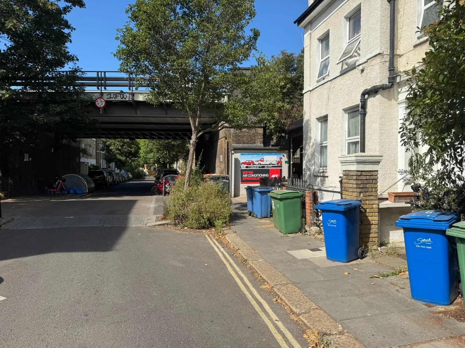 A quiet urban street scene featuring a narrow sidewalk with four trash bins in blue and green, a brick building with white window frames, and an underground overpass with cars parked underneath. There is a sign indicating a height limit of 6 feet 6 inches for the overpass, and a billboard advertising an air conditioning business. The street is lined with trees and parked cars under a bright blue sky.