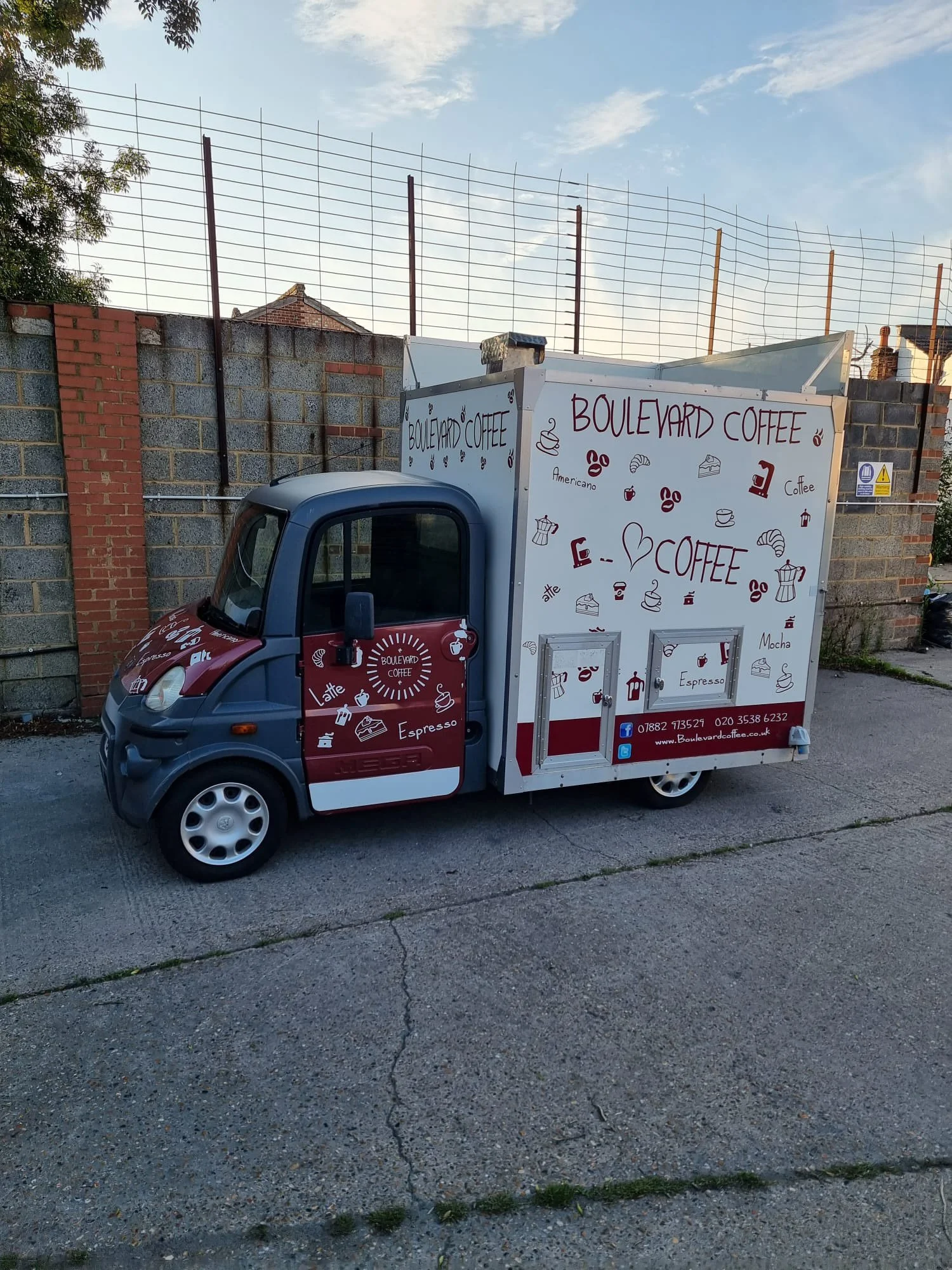 A small blue and red coffee truck with a white side panel displaying various coffee-themed illustrations and text, parked on a street against a brick wall and a wire fence with a partly cloudy sky overhead.