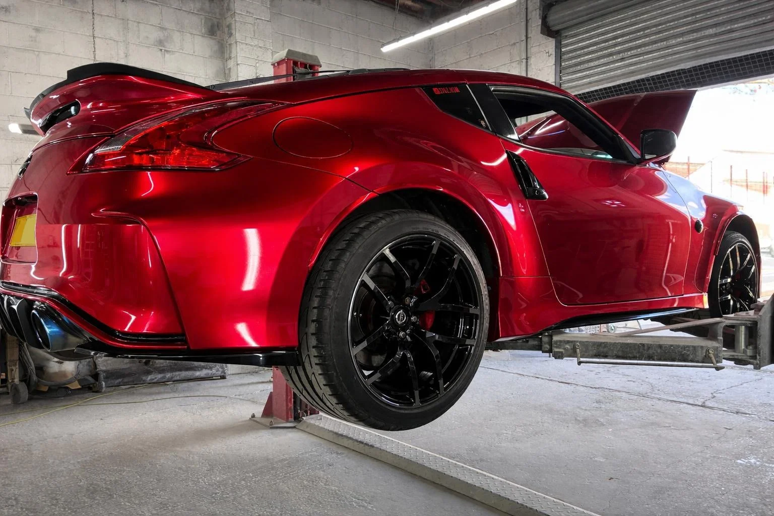 Red sports car on a hydraulic lift inside a garage with an open hood and black wheels.