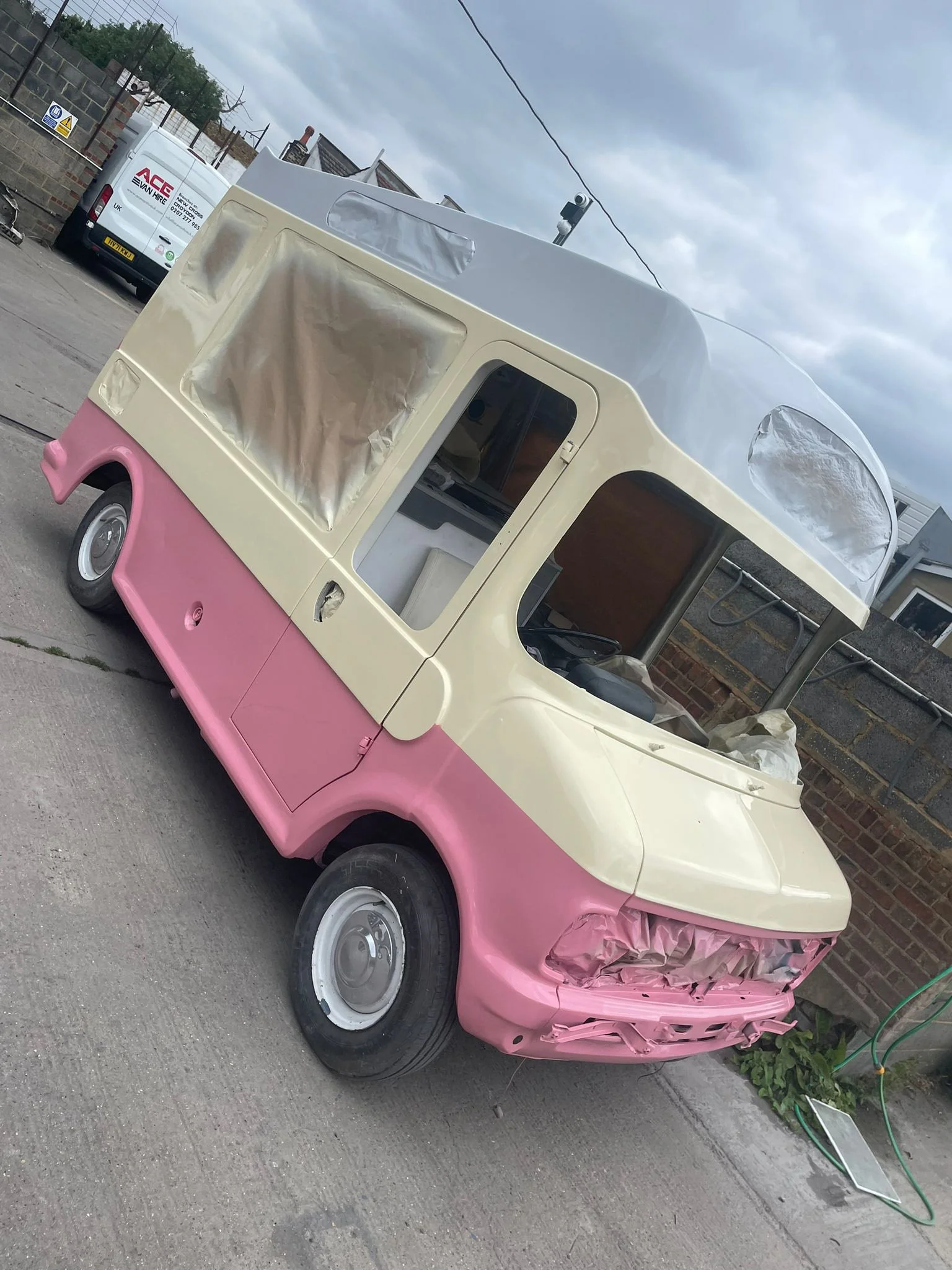 A partially painted vintage van with pink lower half and cream upper half, parked on a driveway with a cloudy sky overhead. The van's windows are covered with protective plastic, and there is a garden hose and some equipment nearby.