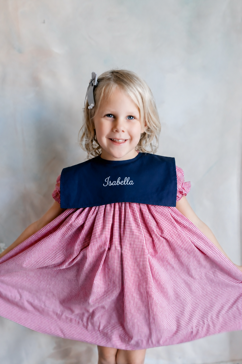 A young girl with blonde hair, blue eyes, wearing a pink and white checkered dress with a navy blue collar embroidered with the name "Isabella," and a bow in her hair, smiling and holding out the skirt of her dress.