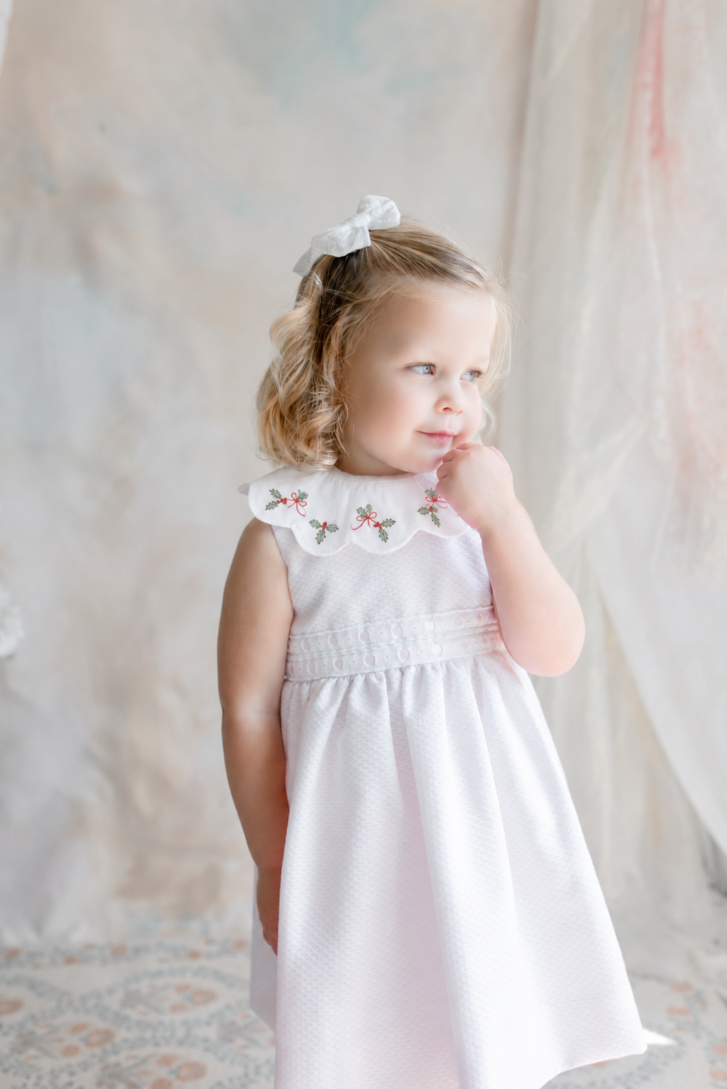 A young girl with blonde, curly hair tied with a white bow, standing near a window with her hand near her chin. She is wearing a white dress with a decorative collar featuring holly and red bow embroidery.