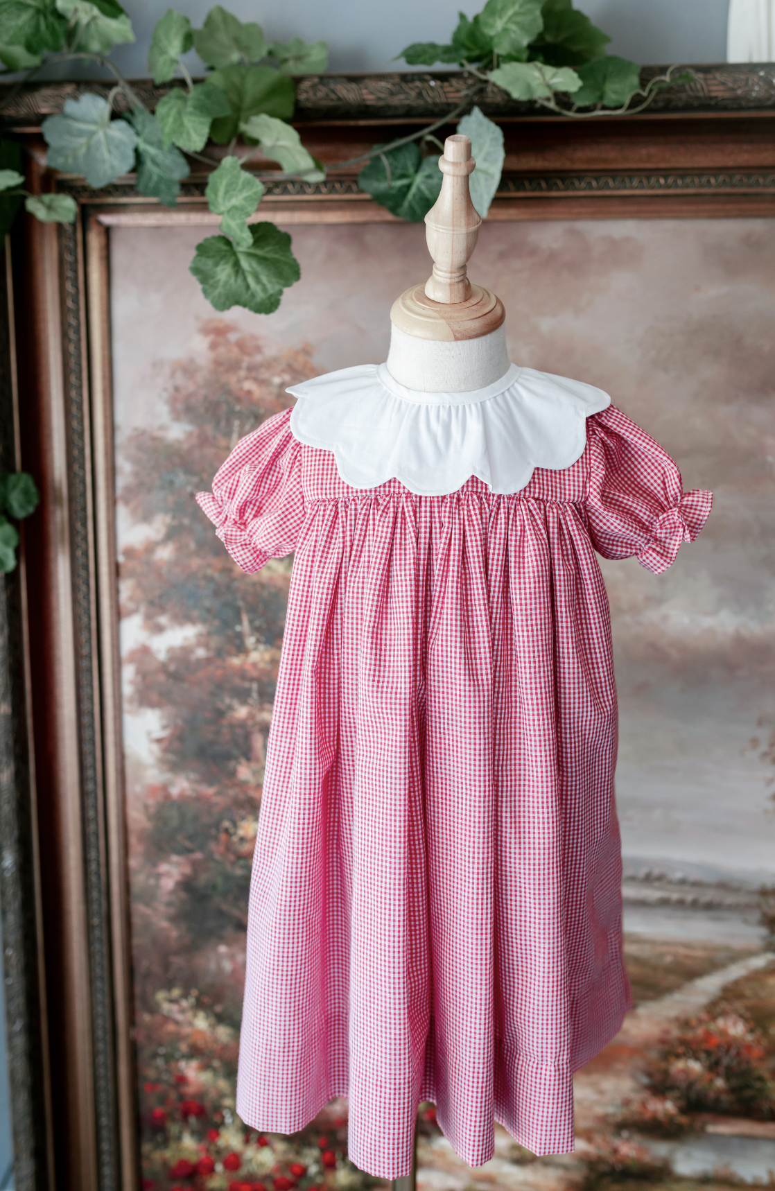 A child's dress displayed on a mannequin with a wooden top, featuring a red and white gingham pattern, a white collar with scalloped edges, and puffy sleeves tied at the ends. Green leaves and a background with autumn-colored trees are visible.