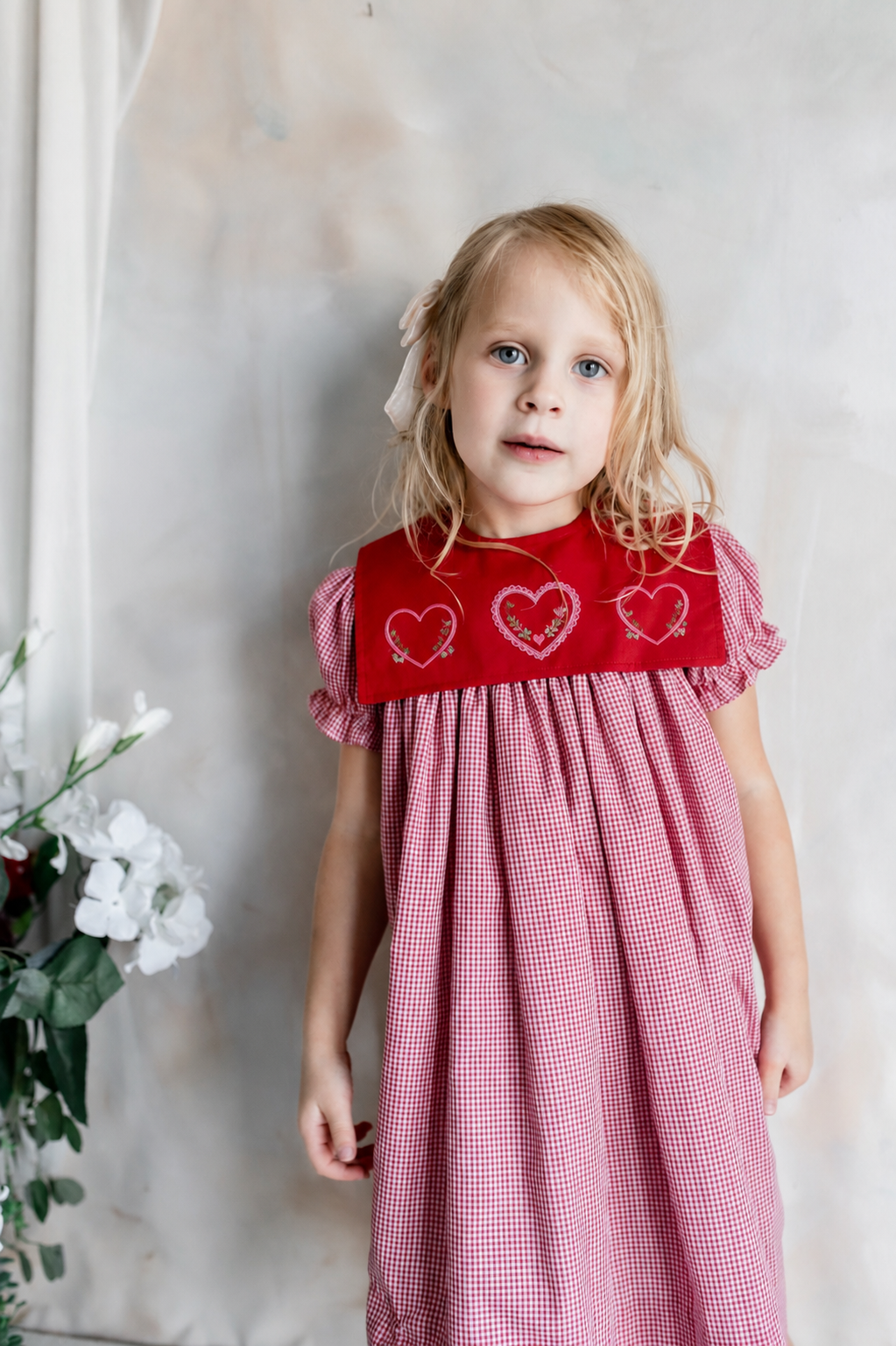 Young girl with blonde hair and blue eyes wearing a red and pink gingham dress with a red collar decorated with embroidered hearts, standing beside white flowers against a light-colored background.