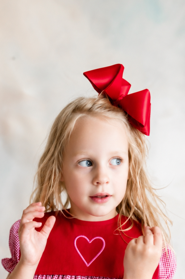 A young girl with blonde curly hair wearing a red dress with a pink heart on the front and a large red bow in her hair, looking to her right.