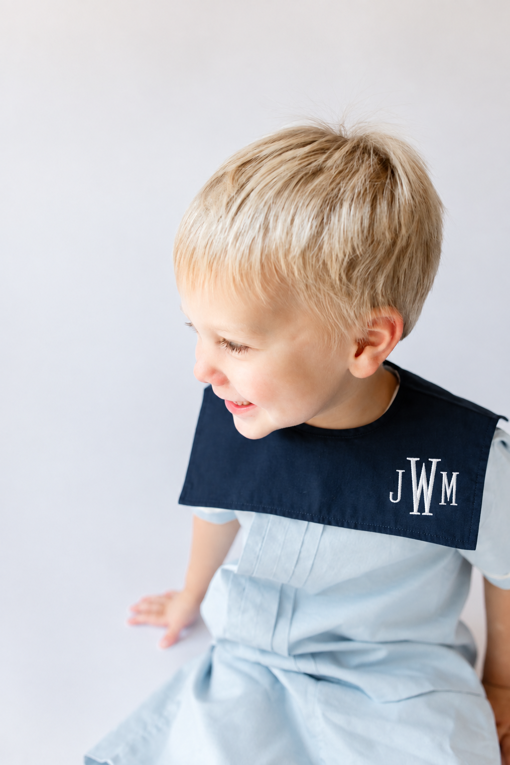 A young boy with blonde hair, smiling and looking down, wearing a light blue smock and a dark blue collar with the embroidered initials JWM.