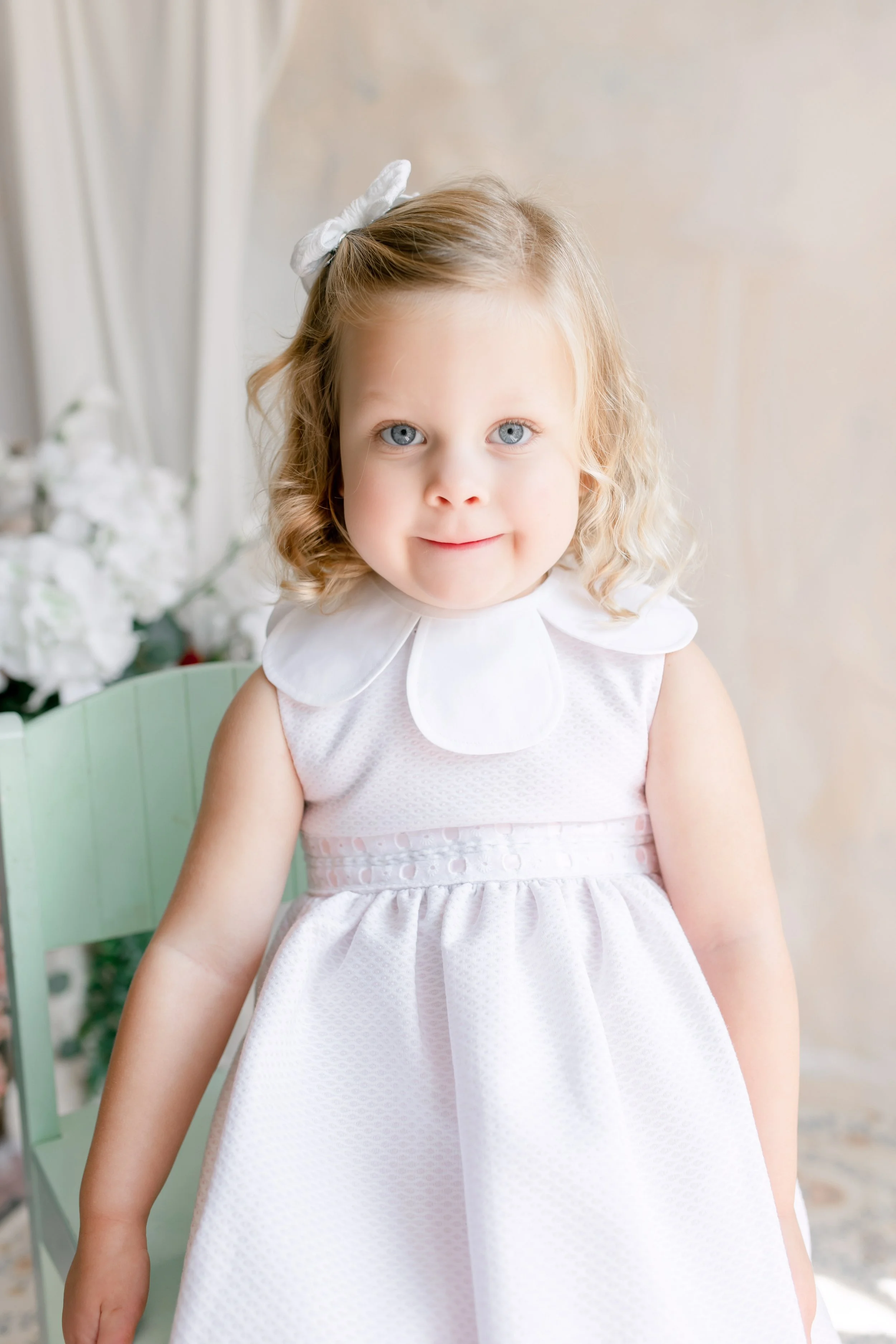 A young girl with curly blonde hair and blue eyes, wearing a white dress with a scalloped collar and a white bow in her hair, sitting on a light green chair with white flowers in the background.