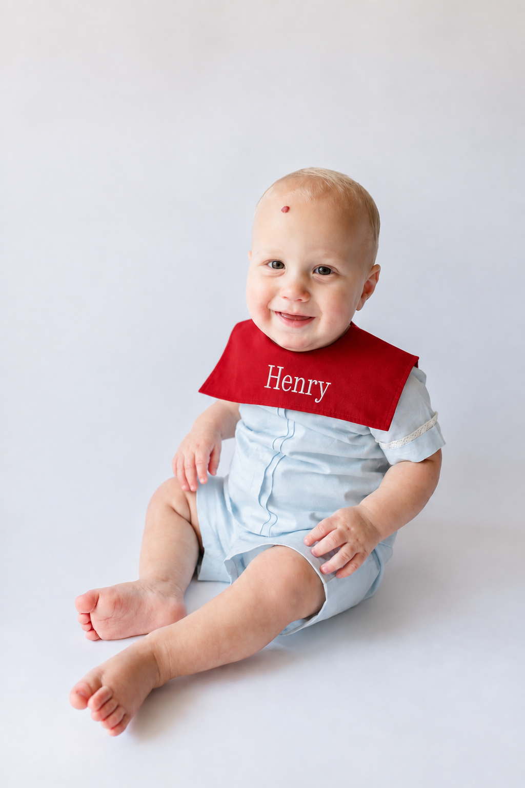 Smiling baby boy with a red decoration on his forehead, wearing a white outfit and a red bib with the name "Henry," sitting on a white surface against a light background.