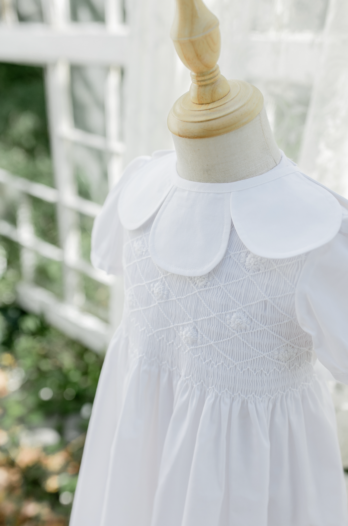 White dress with embroidered floral details and a Peter Pan collar on a mannequin, outdoors with a blurred white fence and greenery in the background.