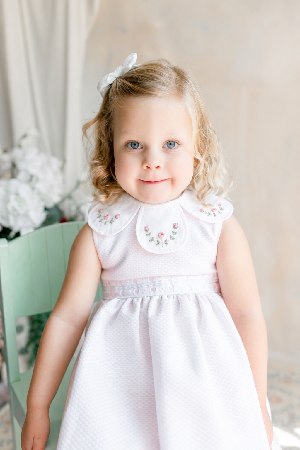 A young girl with blonde curly hair, blue eyes, and a white bow, wearing a white dress with embroidered floral details on the collar, standing indoors with a soft background of flowers and neutral walls.