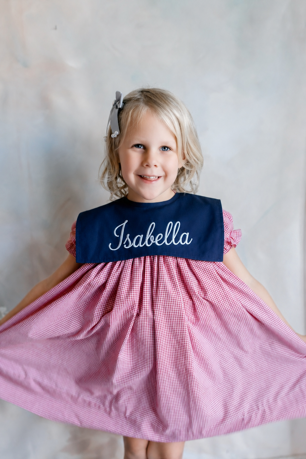 Young girl with blonde hair and a big smile wearing a pink checkered dress and a navy blue sash with the name "Isabella" embroidered on it.