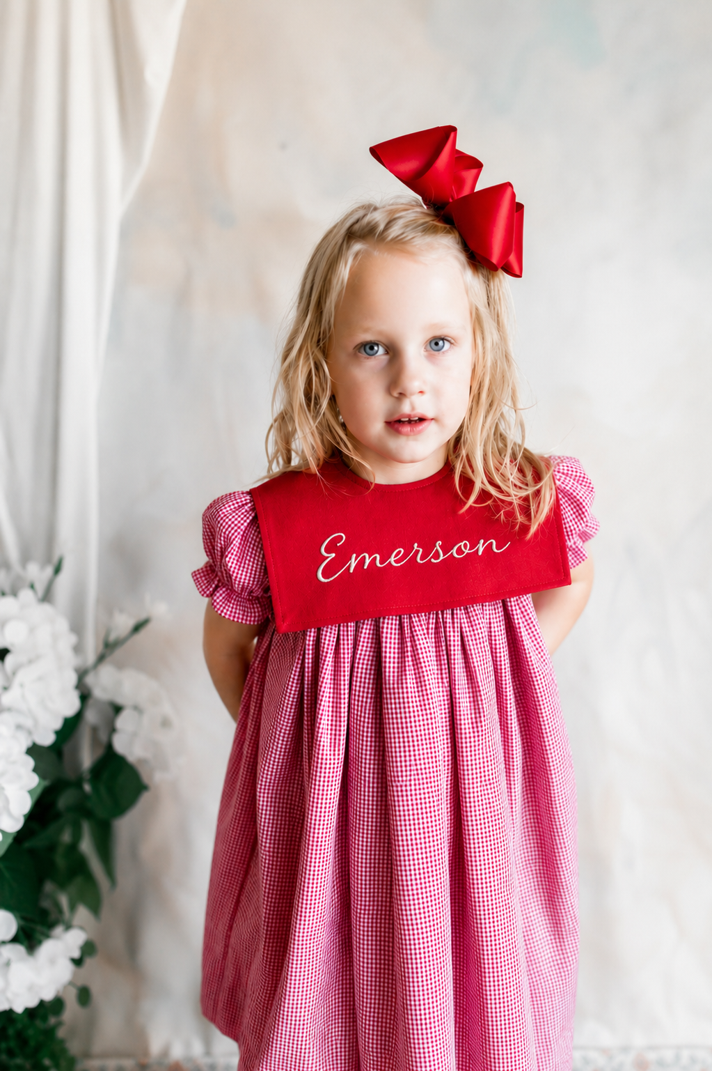 A young girl with blonde, curly hair and blue eyes looking at the camera. She is wearing a red and white checkered dress with puffed short sleeves, a red collar with the name 'Emerson' embroidered on it, and a large red bow on her head. She is standi