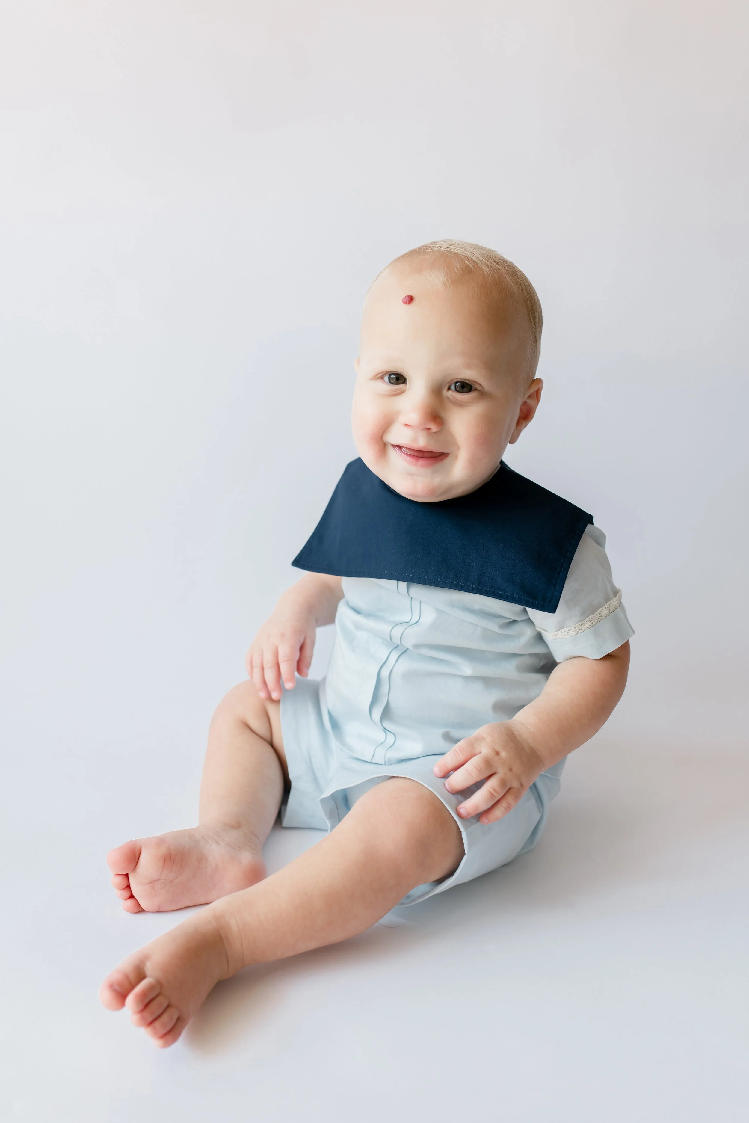 Young smiling child with blond hair sitting on a white surface, wearing a light-colored outfit and a navy blue bib, with a small red mark on their forehead, against a plain light background.