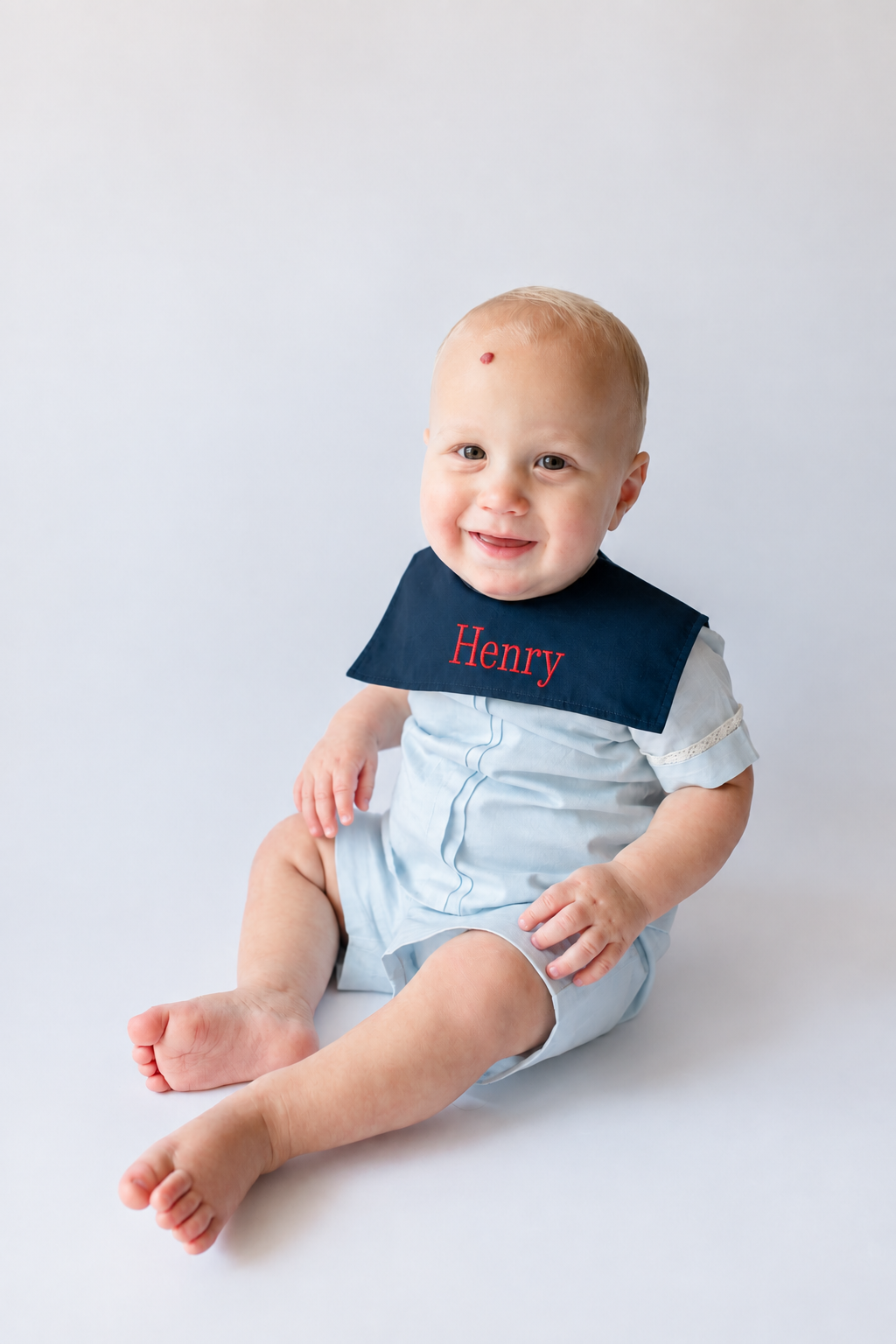 Smiling baby boy wearing a blue bib with 'Henry' embroidered in red, sitting on a white background, with a small red mark on his forehead.
