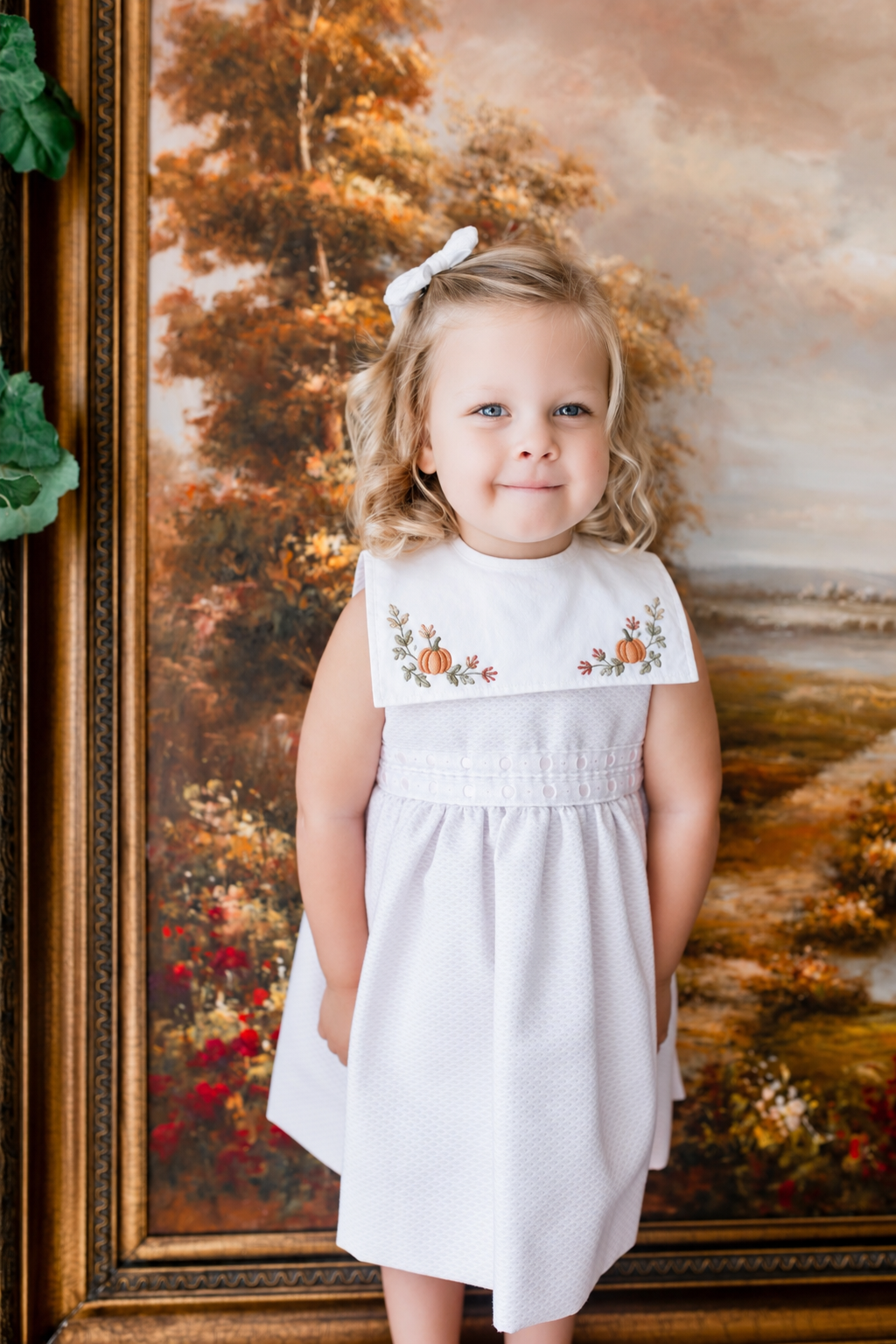 A young girl with blonde curly hair and a white dress with embroidered pumpkins stands in front of an autumn landscape painting. She has a white bow in her hair and is smiling softly.