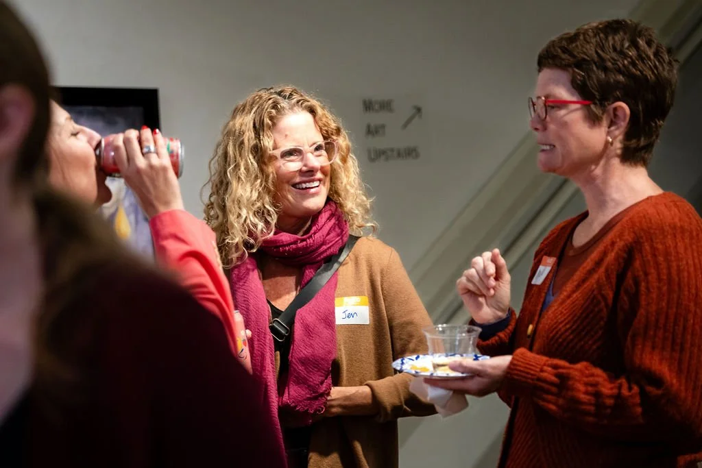 One woman with blond curly hair wearing a pink scarf and brown sweater smiling at a woman wearing a red sweater and glasses who is speaking. Another woman, mostly out of the frame, is holding a can up to drink.