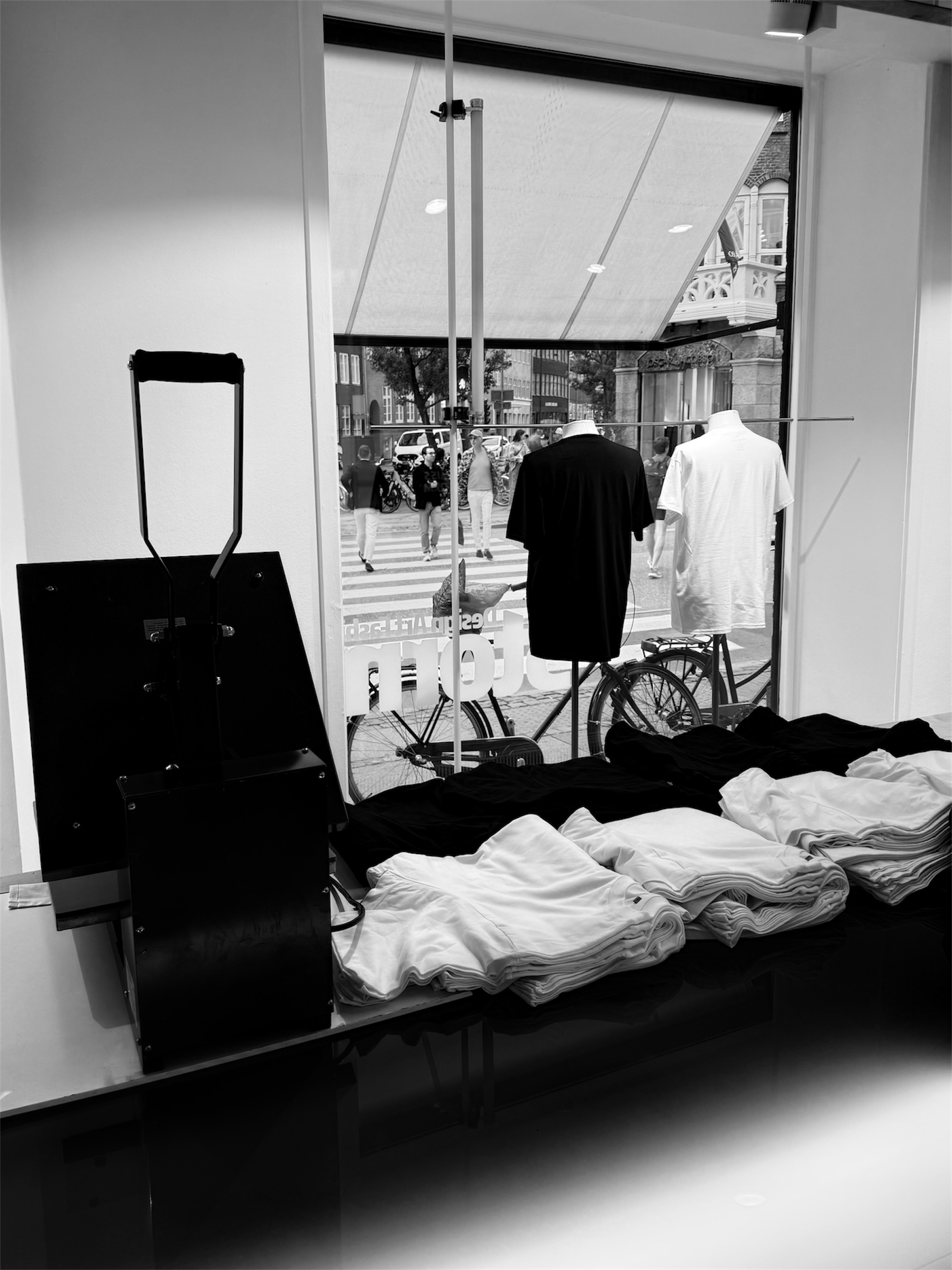 The image shows the interior of a store with clothing items, specifically folded shirts on a table, and two mannequins dressed in clothing, one black and one white, standing by the window. Outside the window, pedestrians are crossing the street, with cars parked along the street.