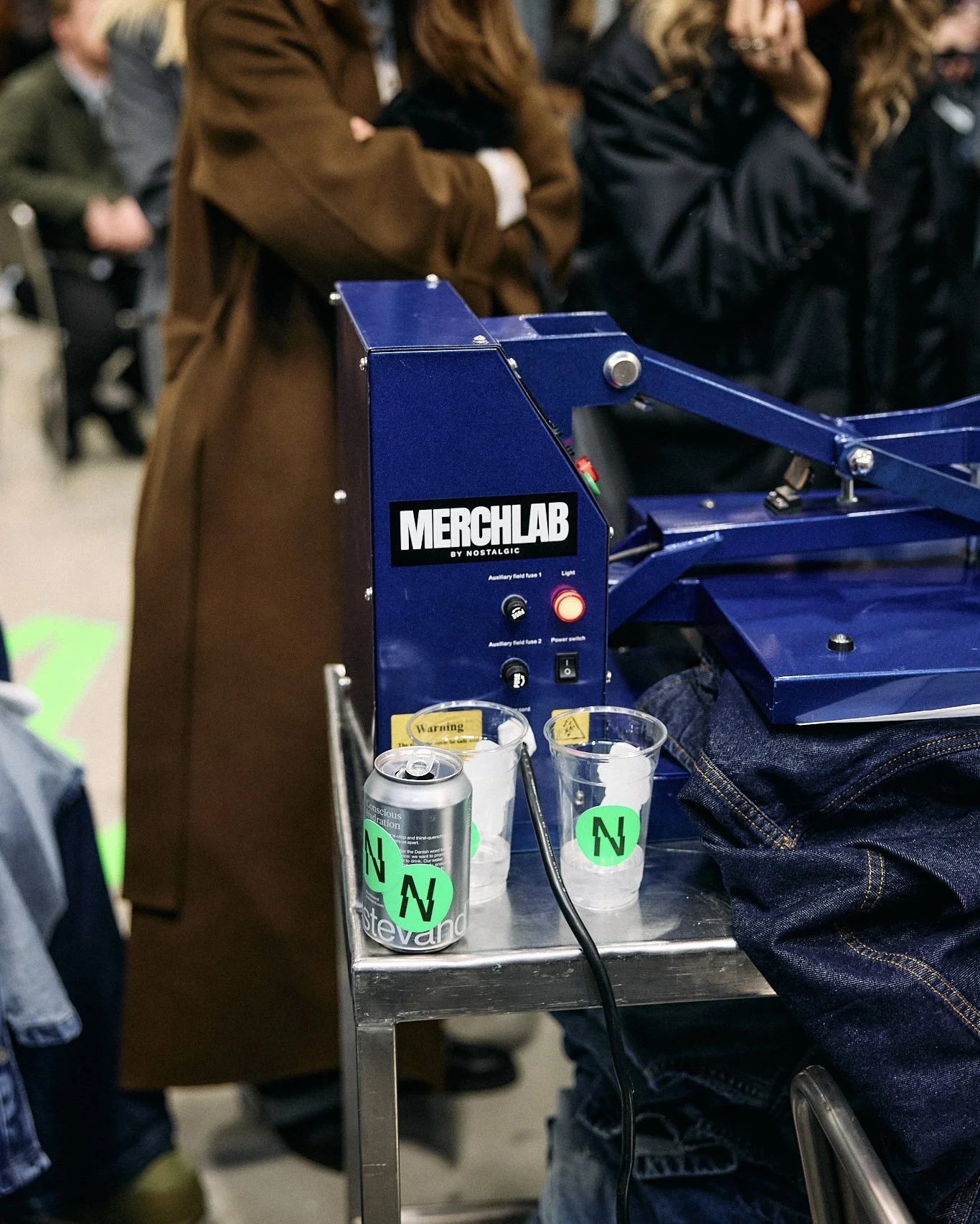 Person pressing a heat press onto a grey t-shirt at a live event, with other people and equipment visible nearby.