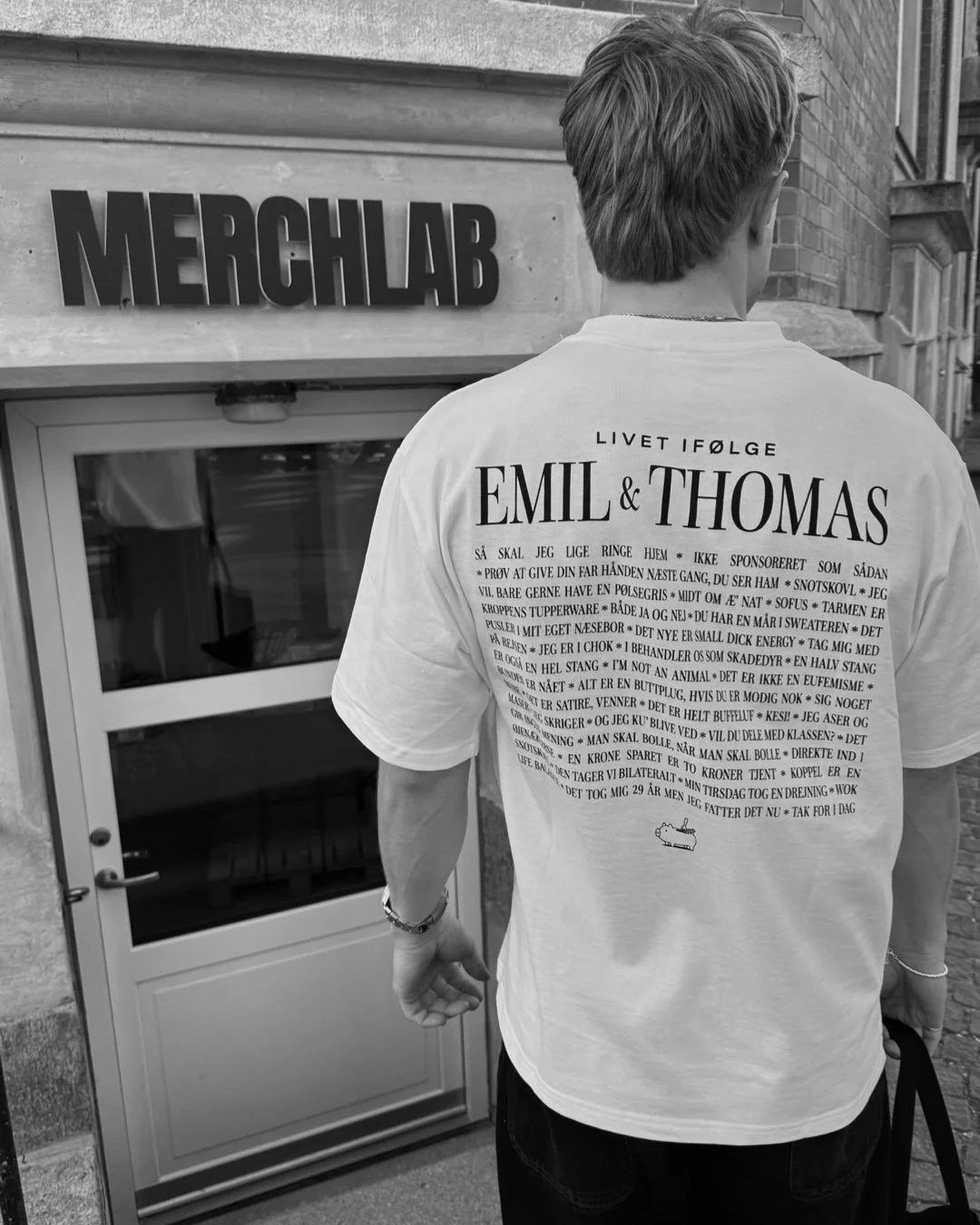 A person with short hair wearing a white T-shirt with text in Danish on the back, standing in front of a door with a sign that says 'MERCHLAB.'