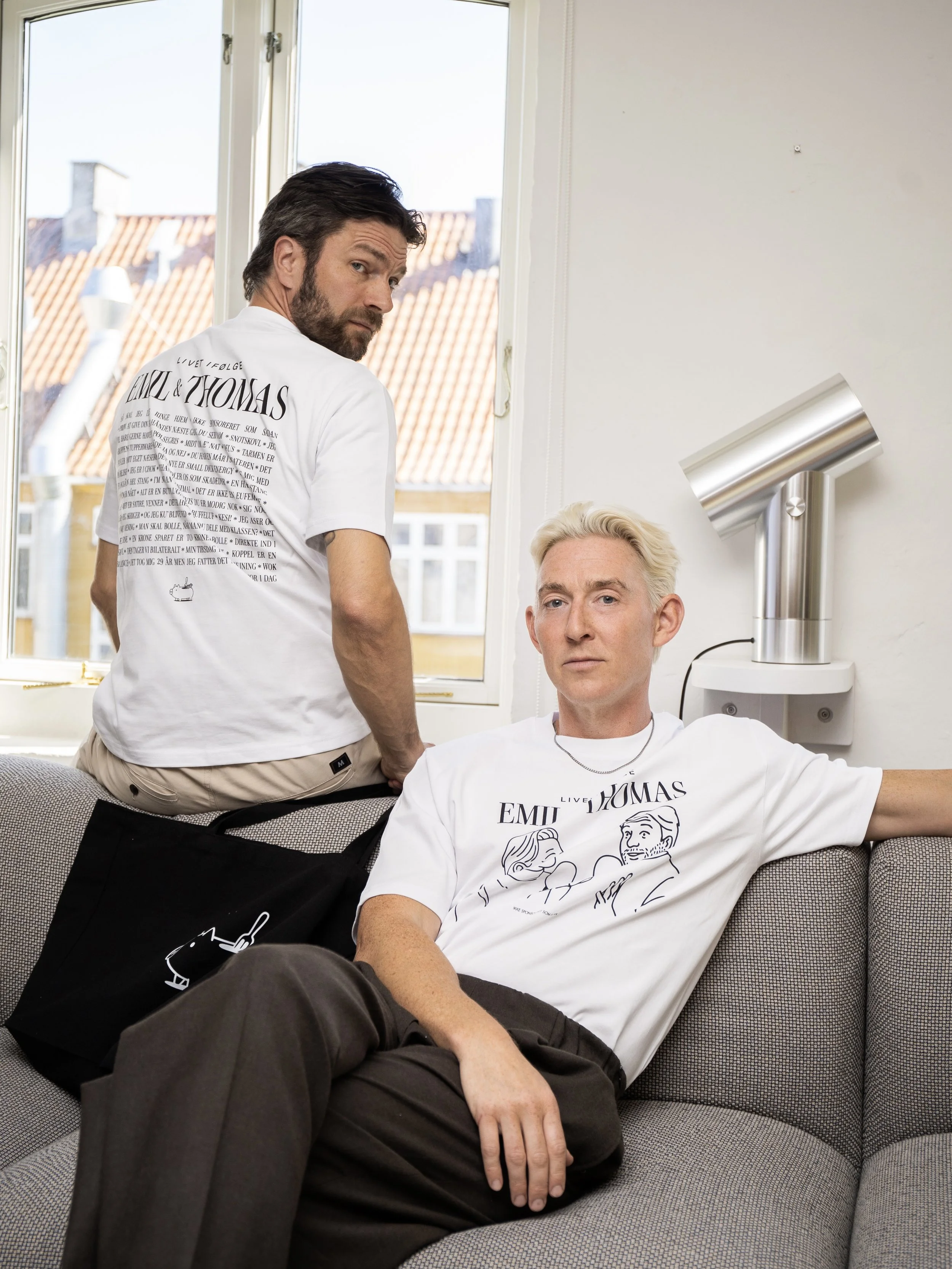 Thomas Skov and Emil Thorup. Both are wearing white T-shirts with graphic and text. The man standing looks over his shoulder at the camera, and the man sitting appears relaxed, with his eyes open and head resting against the couch.