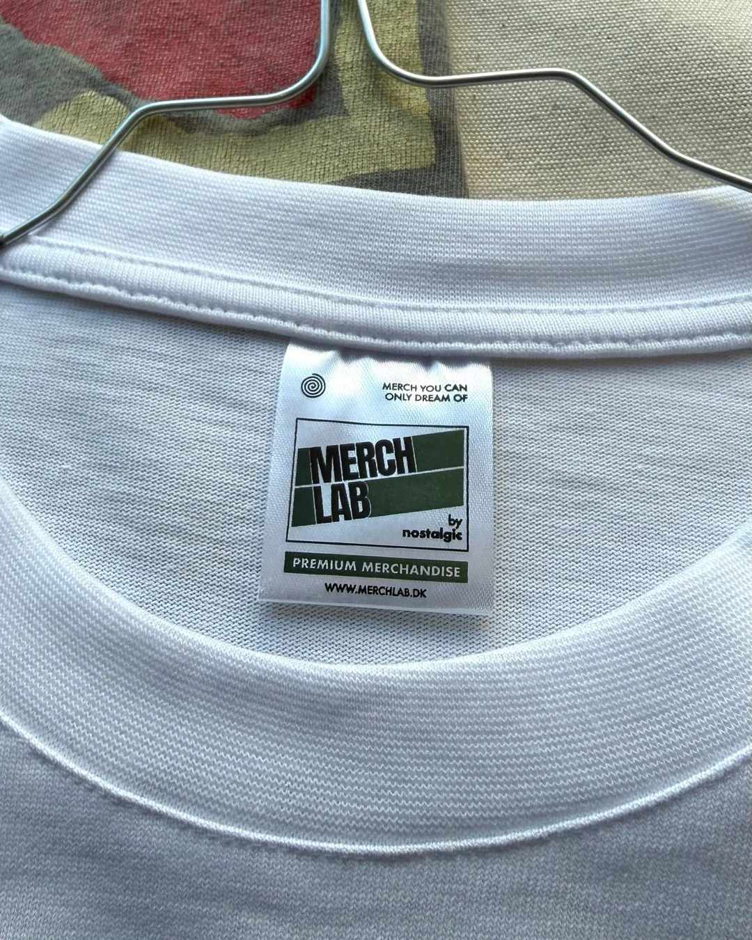 Close-up of a white T-shirt on a hanger with a tag that says 'MERCH LAB by nostalgic, premium merchandise, www.merchlab.dk'