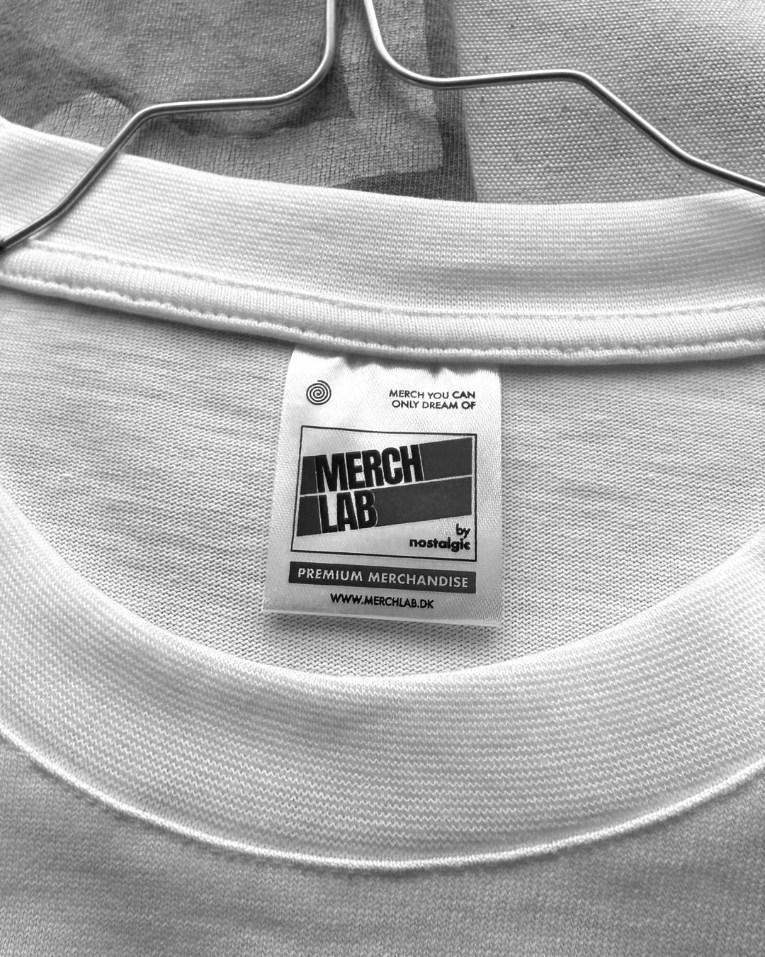 Close-up of a white T-shirt on a hanger with a label from 'Merch Lab' by Nostalgic, featuring text about premium merchandise and a website URL.