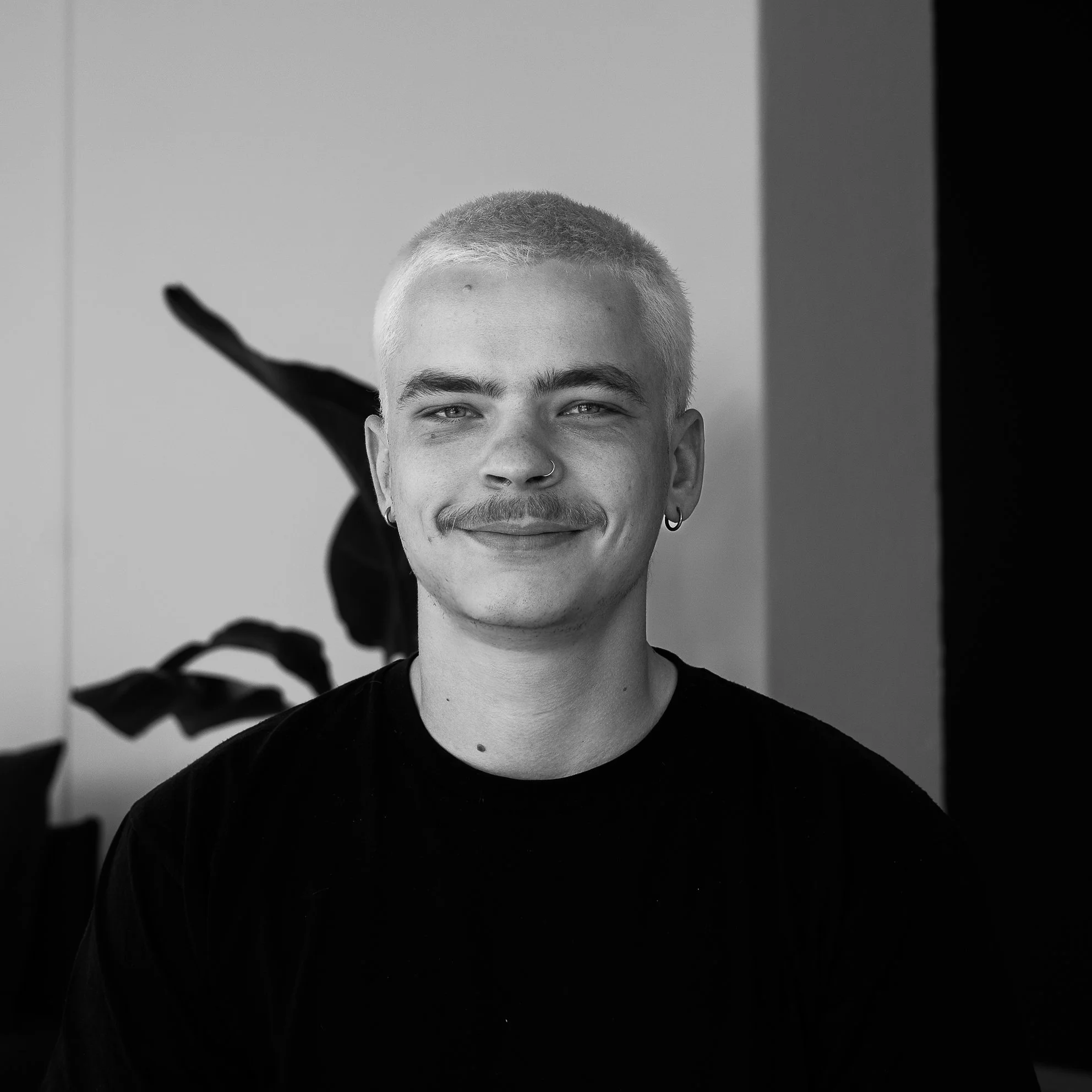Portrait en noir et blanc d'une personne avec une coupe courte, souriante, portant un t-shirt noir, dans un intérieur avec un fond minimaliste et une plante visible.
