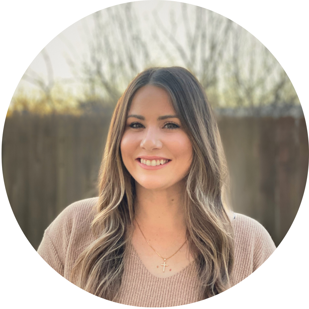 A smiling woman with long wavy hair standing outdoors with a blurred fence and trees in the background.