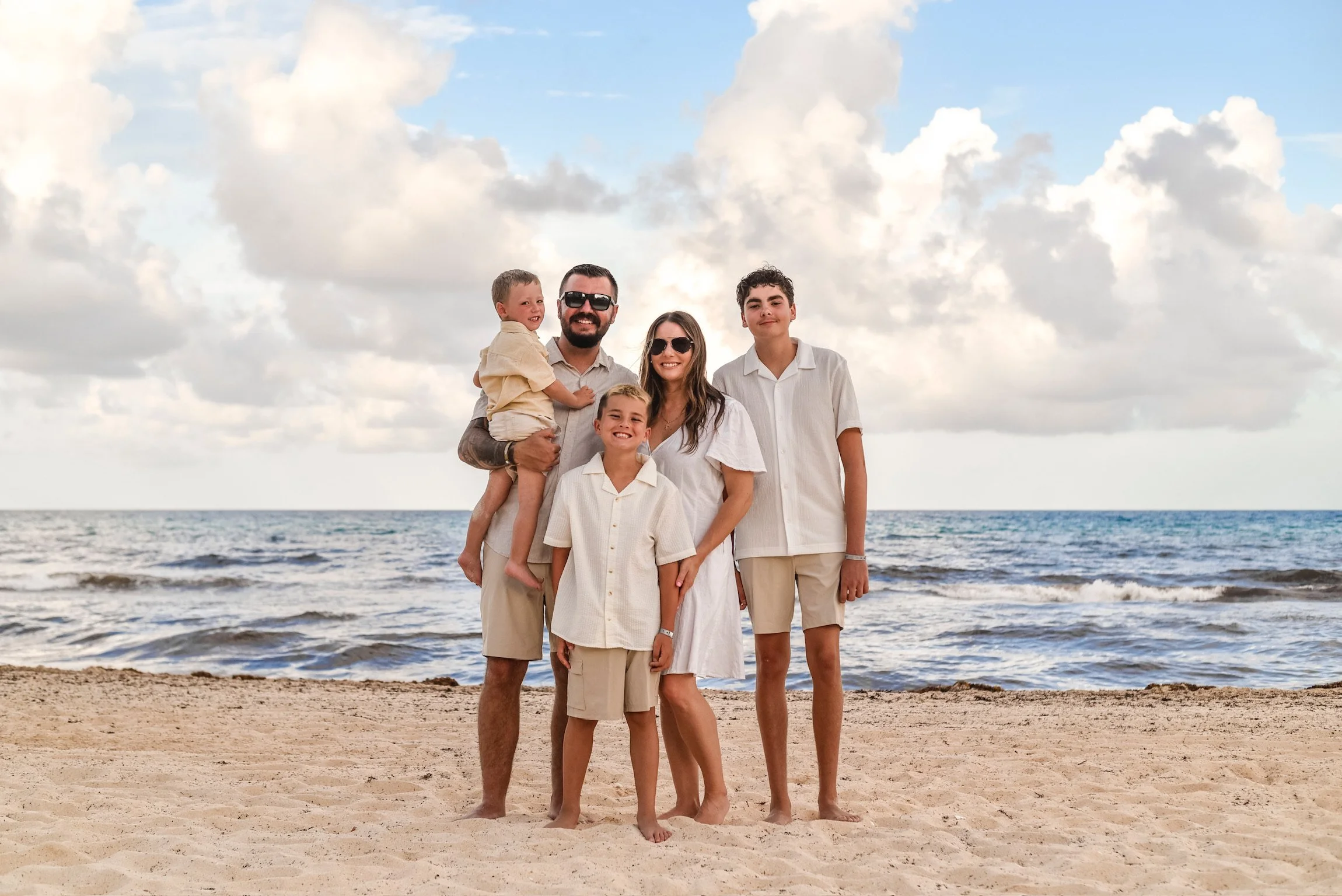 A family of five standing on a sandy beach with ocean waves and a cloudy sky in the background, all dressed in white and beige clothing.