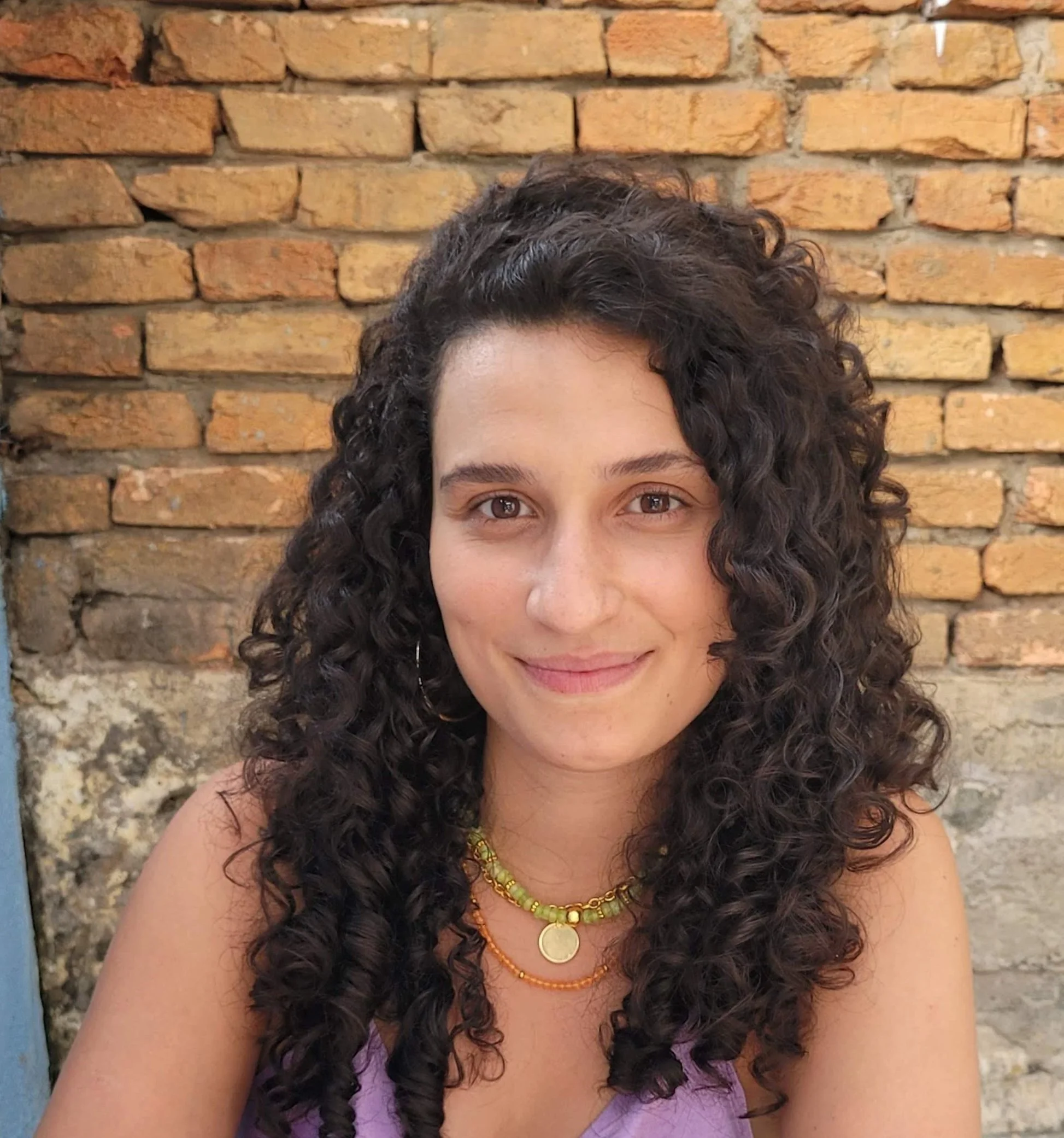 Pessoa com cabelo cacheado sorrindo em frente a uma parede de tijolos.