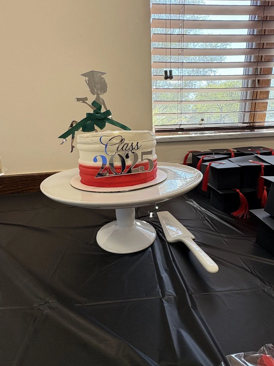 Graduation-themed cake with topper of graduate silhouette and 'Class of 2025' decoration on a white cake stand, on a black tablecloth next to black graduation caps with red tassels, near a window with blinds.