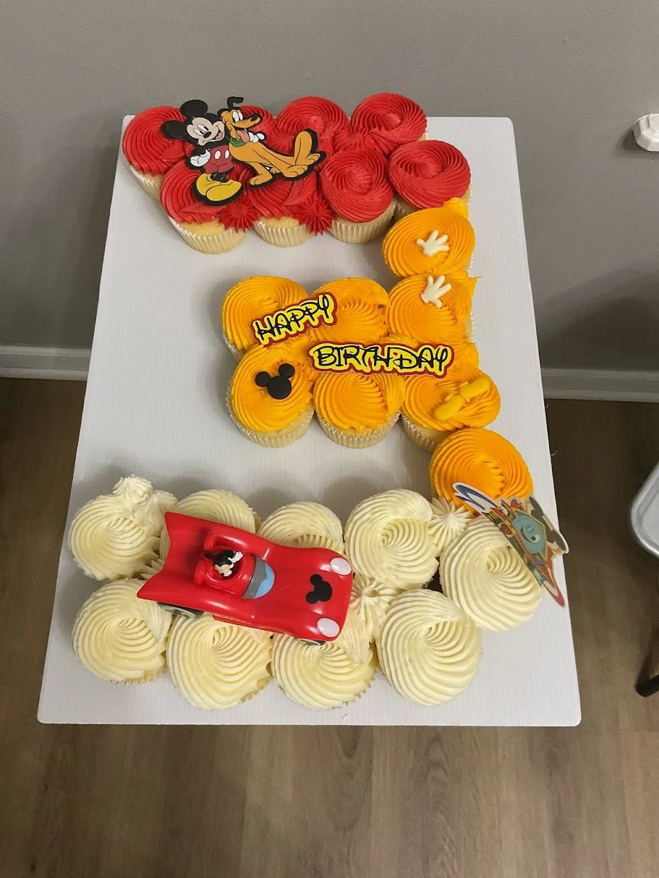 Cupcake cake decorated with Mickey Mouse and Pluto toppers, with a large number 1 shape made of cupcakes, featuring yellow, orange, and white frosting, and birthday messages reading "HAPPY BIRTHDAY."