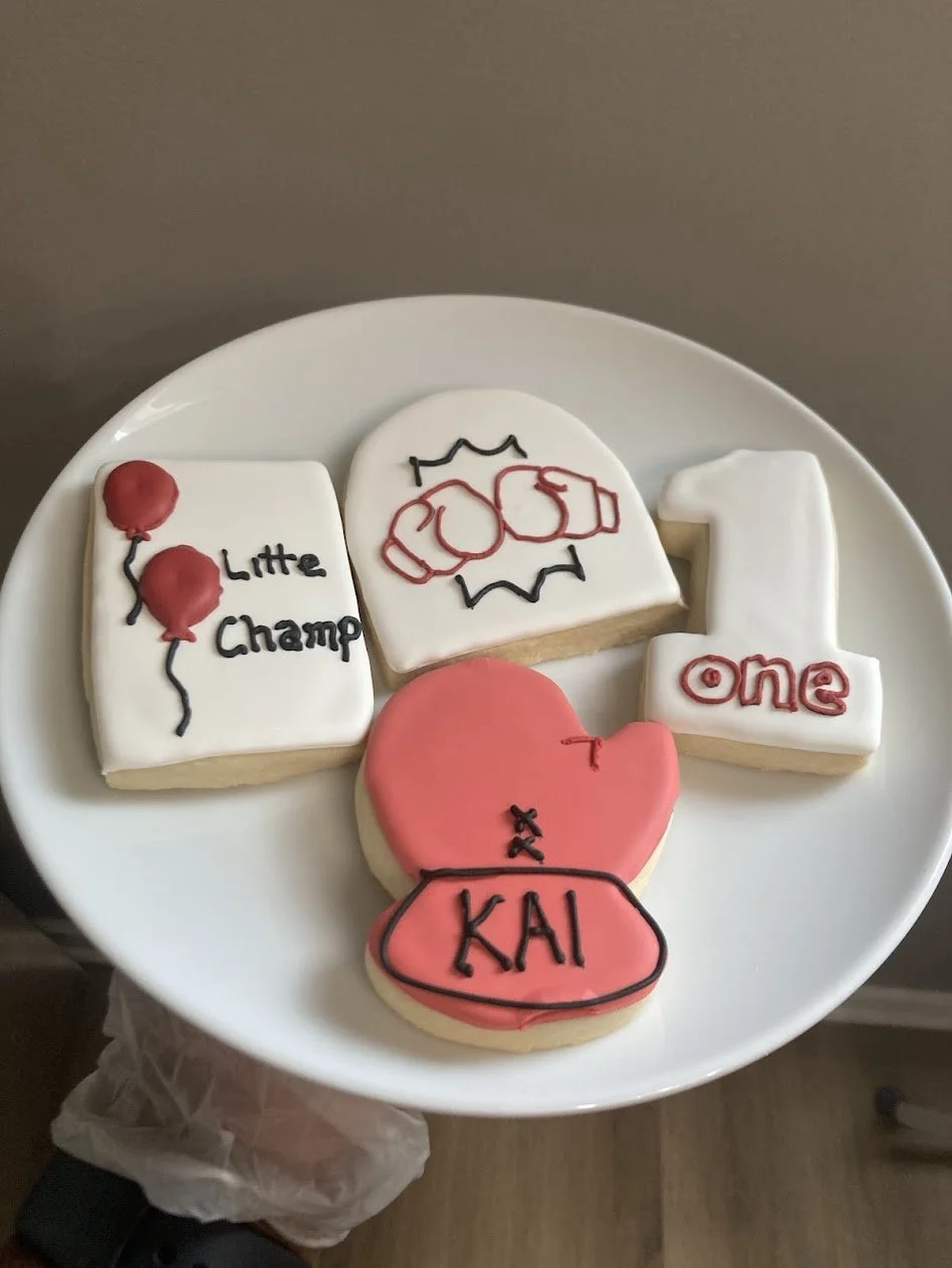 Cookies decorated with birthday themes, including balloons, a crown, a number one, and a heart with initials 'KAI' for a first birthday celebration.