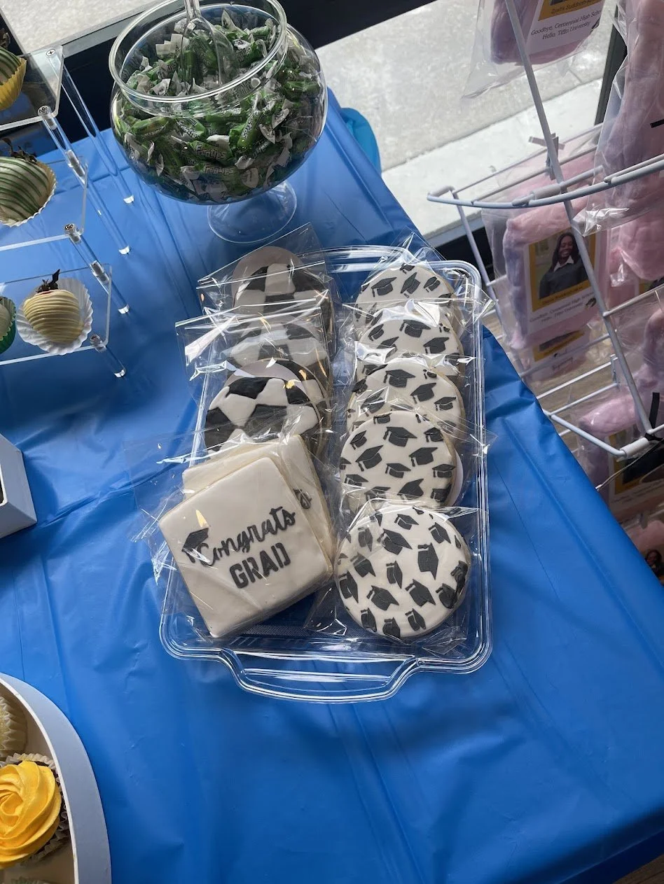 Decorated cookies on a table, including square cookies with 'Congrats Grad' written and round cookies with a graduation cap pattern, all wrapped in plastic. A glass bowl with green and white candies is nearby.