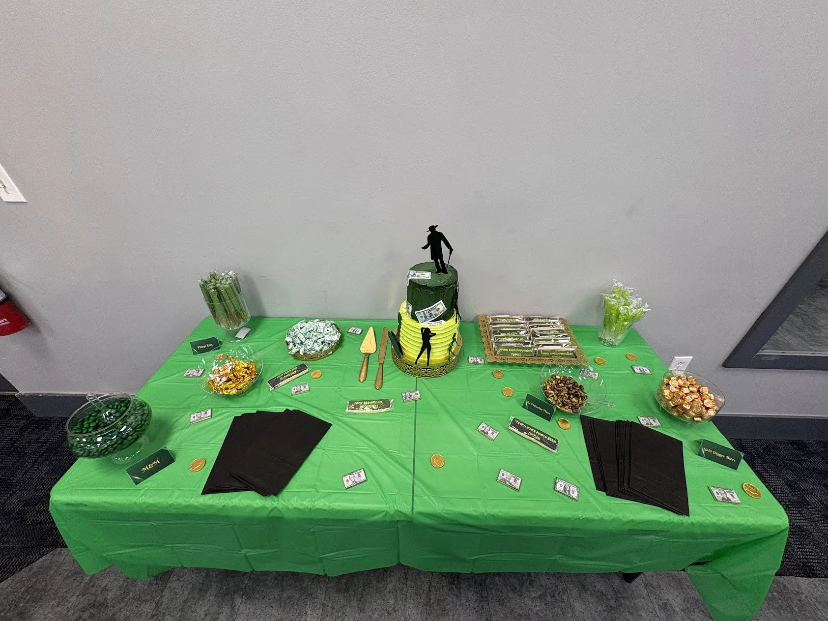 A table decorated in green with various candies and a themed cake, featuring dollar bill decorations and figurines resembling construction workers, likely for a St. Patrick's Day celebration.