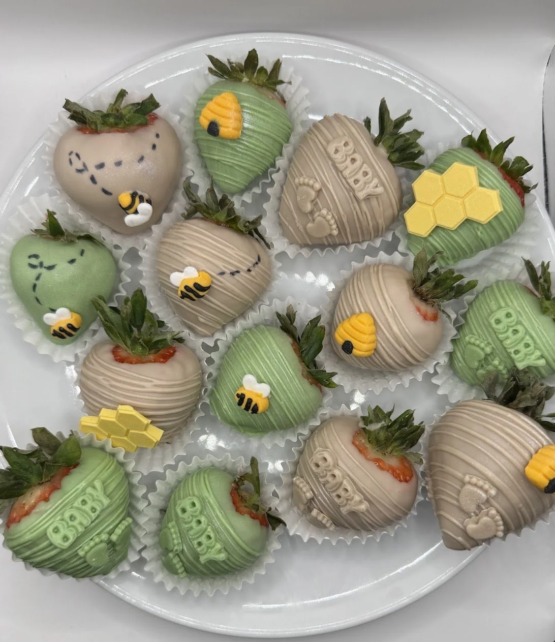 A white tray holding chocolate covered strawberries decorated with bee, honeycomb, and hive designs in yellow, black, white, and beige colors.